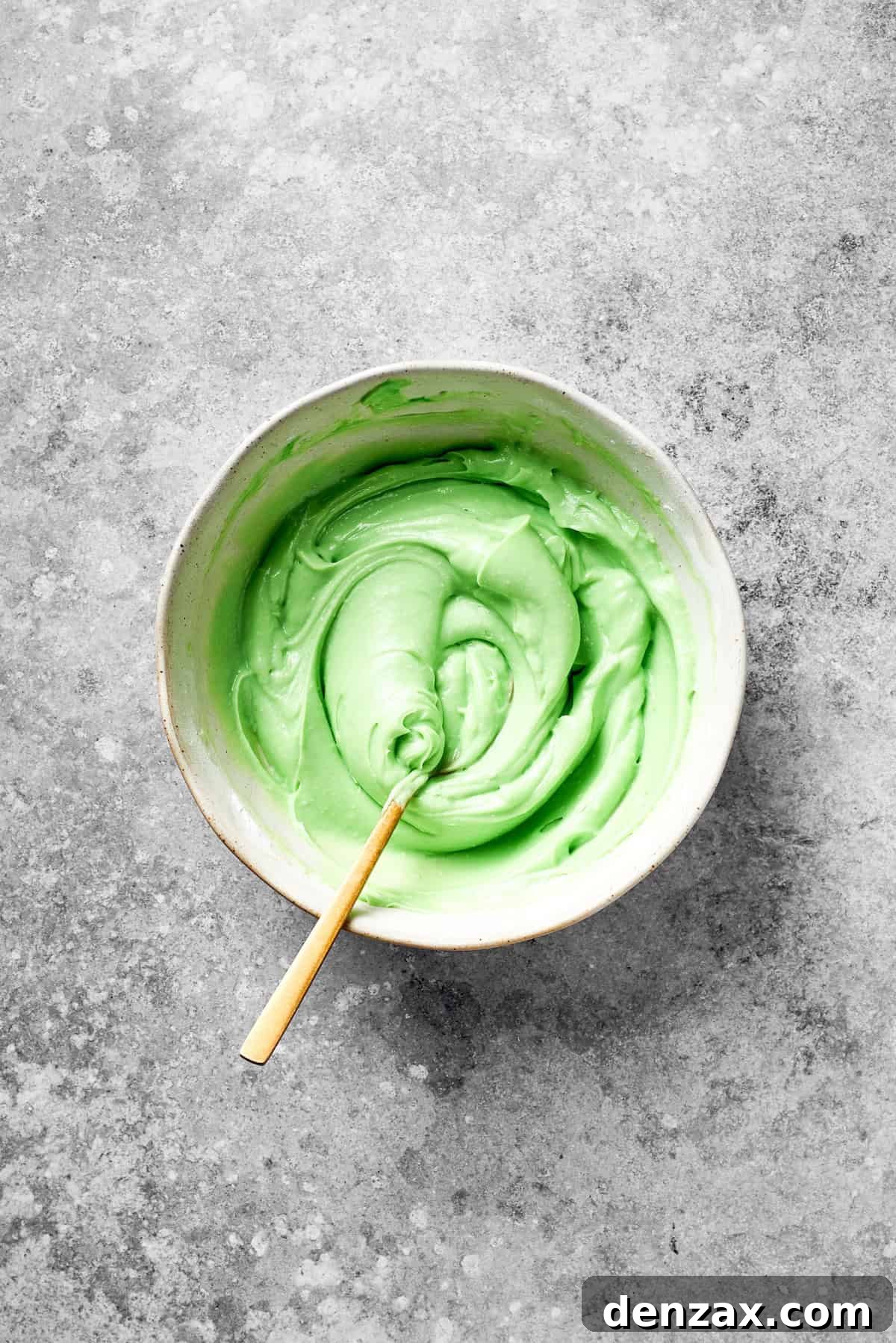 Stir Green Icing Until Smooth for Grinch Cake A spoon actively stirs green food coloring into a bowl of icing, creating a smooth, uniformly colored frosting.