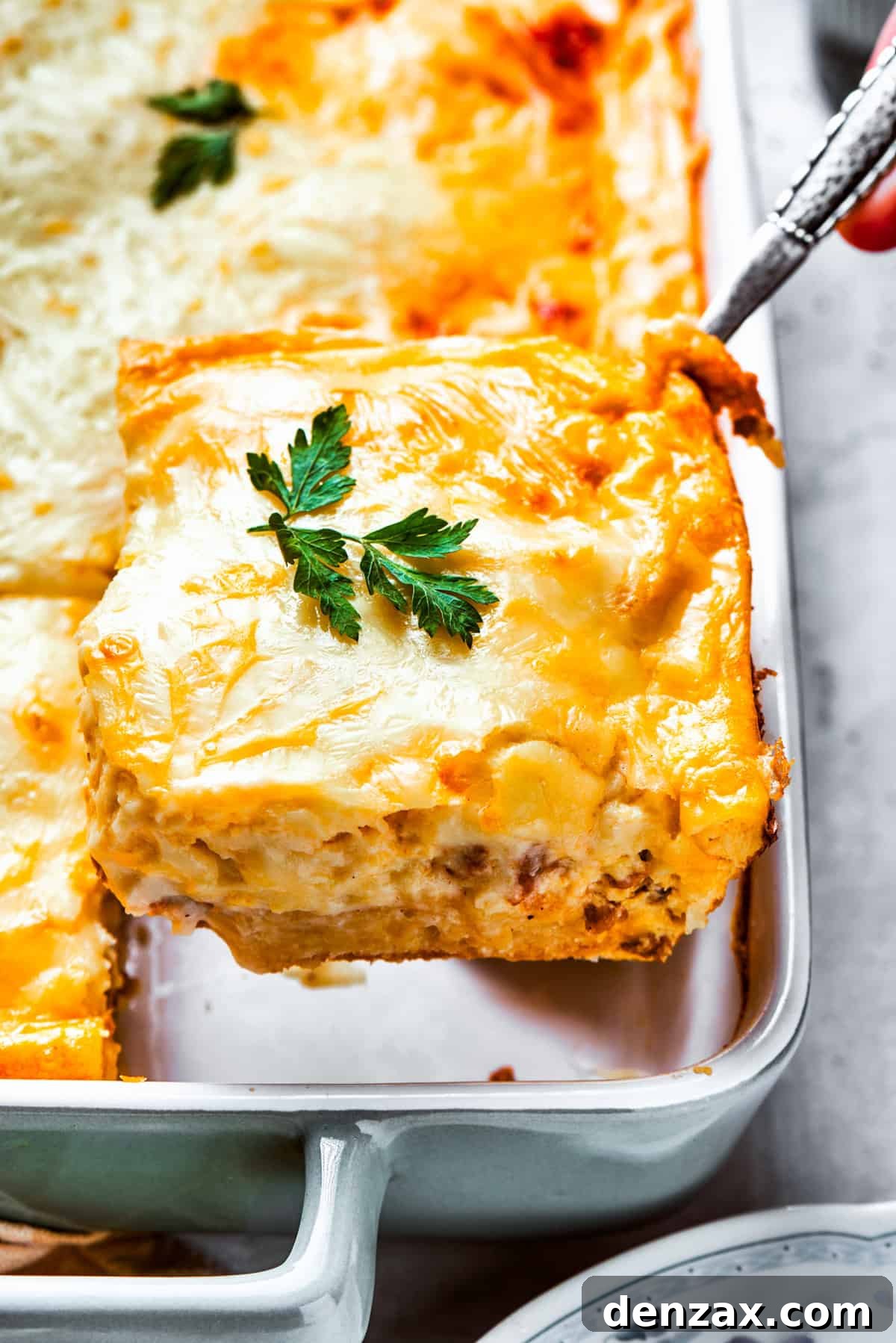 A serving spoon scoops out a generous slice of hot, cheesy biscuits and gravy casserole from the baking dish.