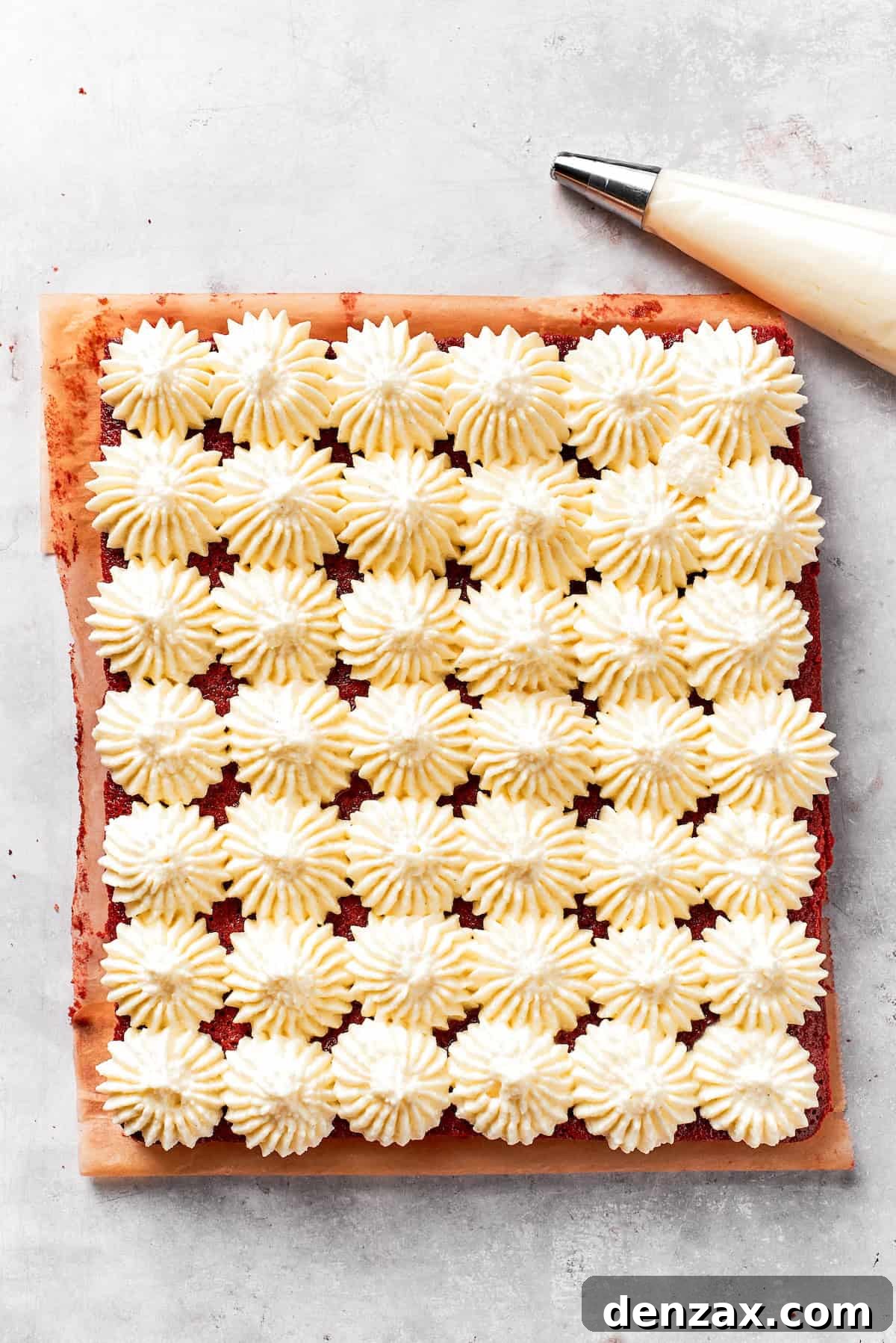 Thick, white cream cheese frosting is being piped in elegant swirls onto a baked sheet of vibrant red velvet brownies.