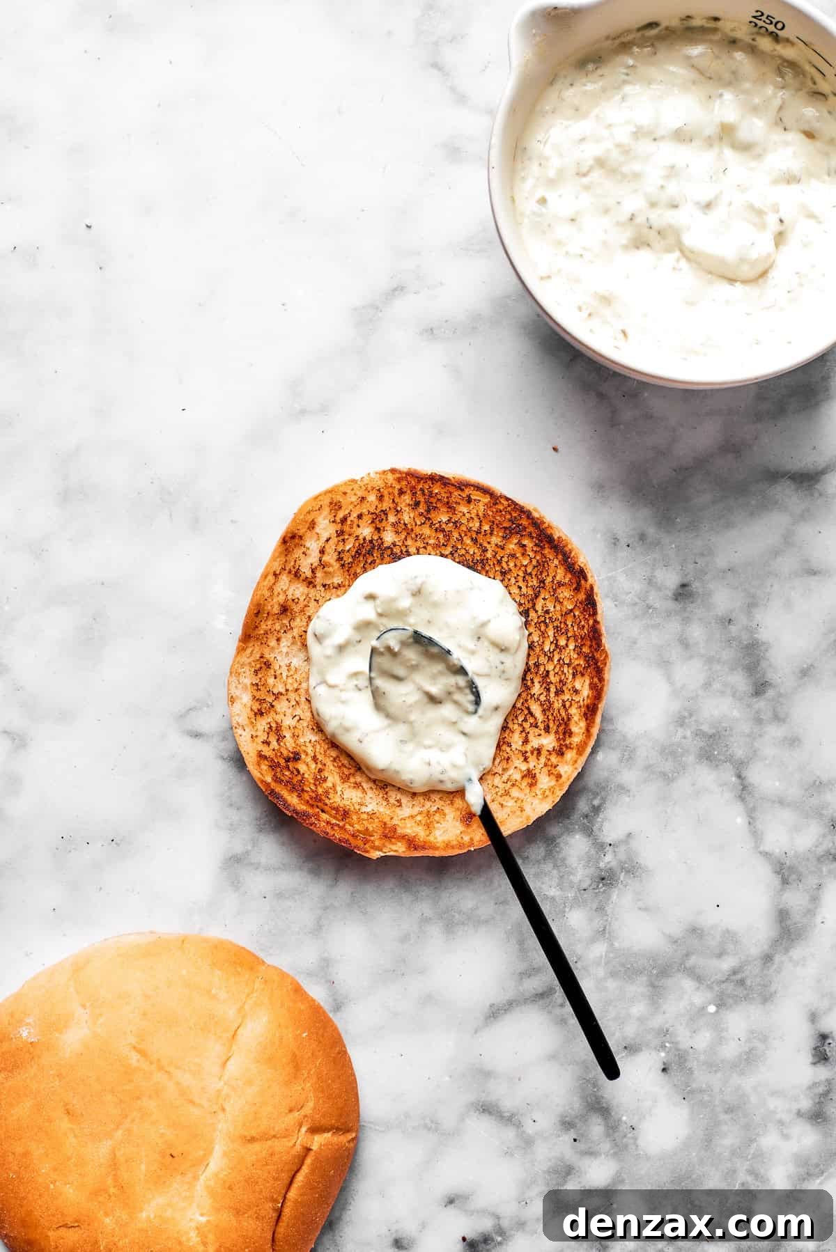 Creamy tartar sauce being generously spread on a golden-toasted brioche bun, preparing it for the fish fillet.
