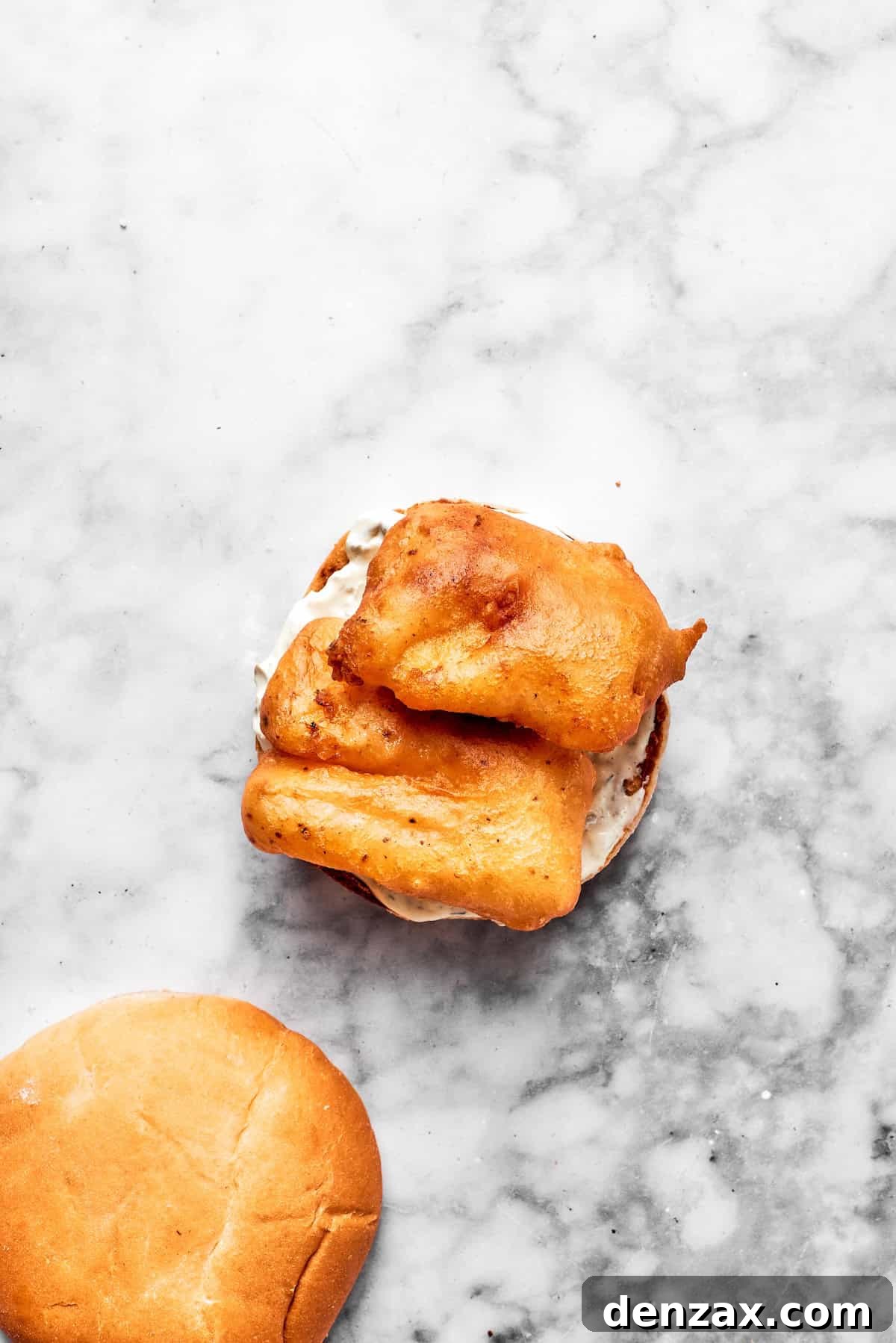 A freshly fried, golden fish fillet being placed onto a toasted bun generously spread with tartar sauce.