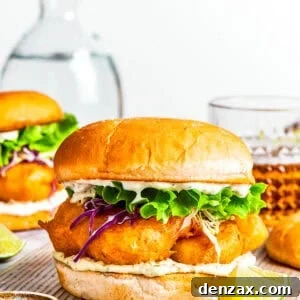 A delicious homemade fish sandwich served on toasted buns with another sandwich and drinks in the background, making for a perfect meal.