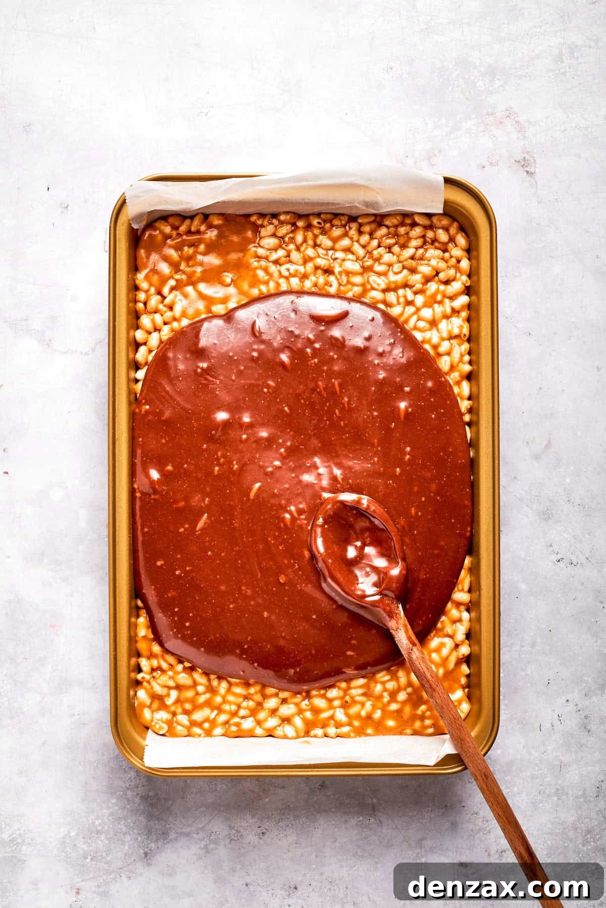 The melted chocolate and butterscotch topping being spread evenly over the crispy rice cereal base in a baking pan.