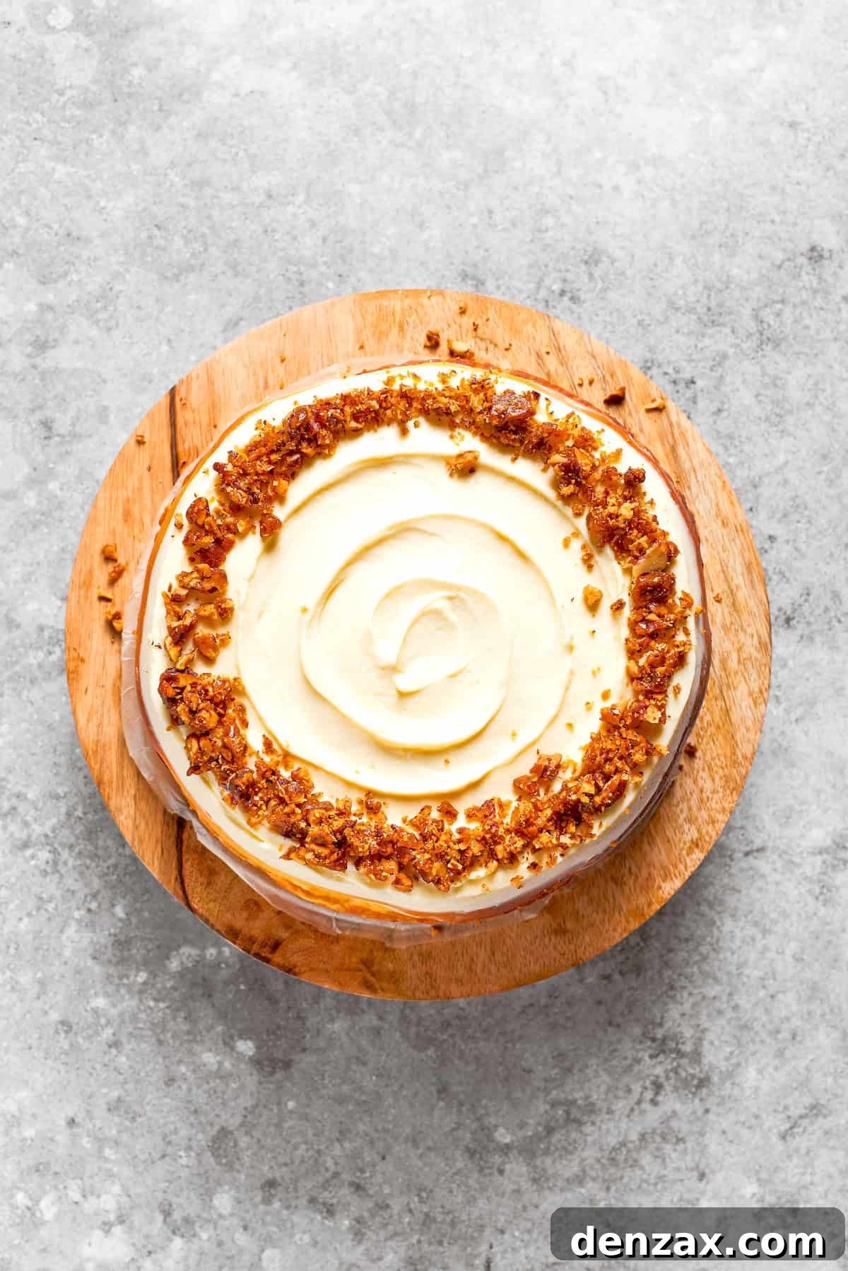 A fully frosted carrot cake cheesecake, topped with caramelized pecans, presented on a wooden cake stand.