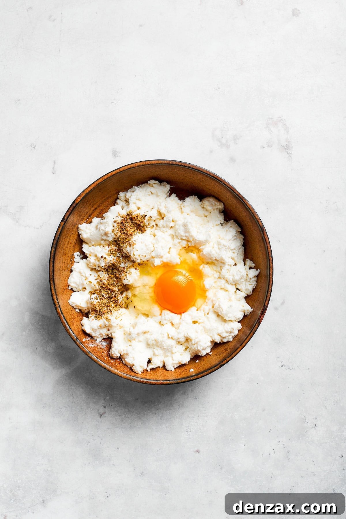 An egg being cracked into a bowl of creamy ricotta cheese.