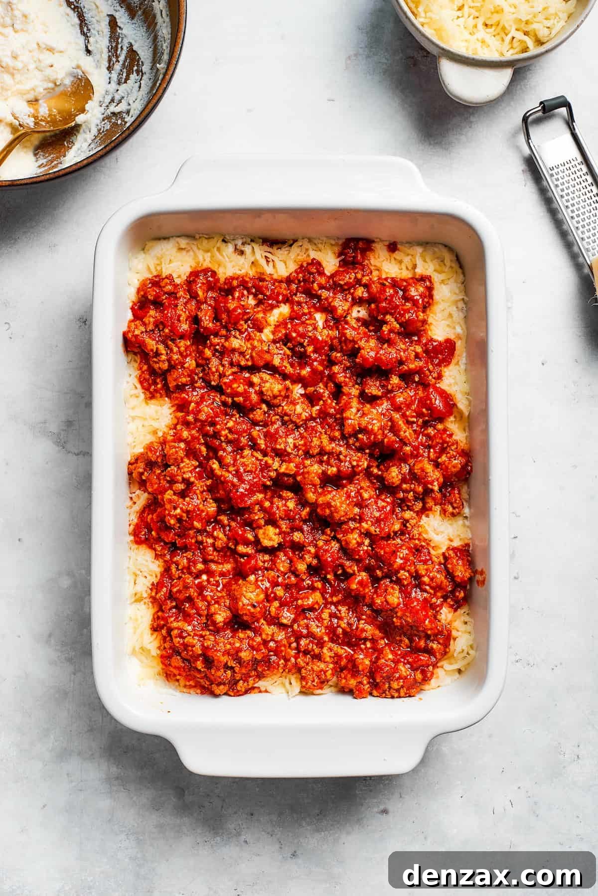 A ladle of rich meat sauce being spread over a cheese layer in the lasagna baking dish.