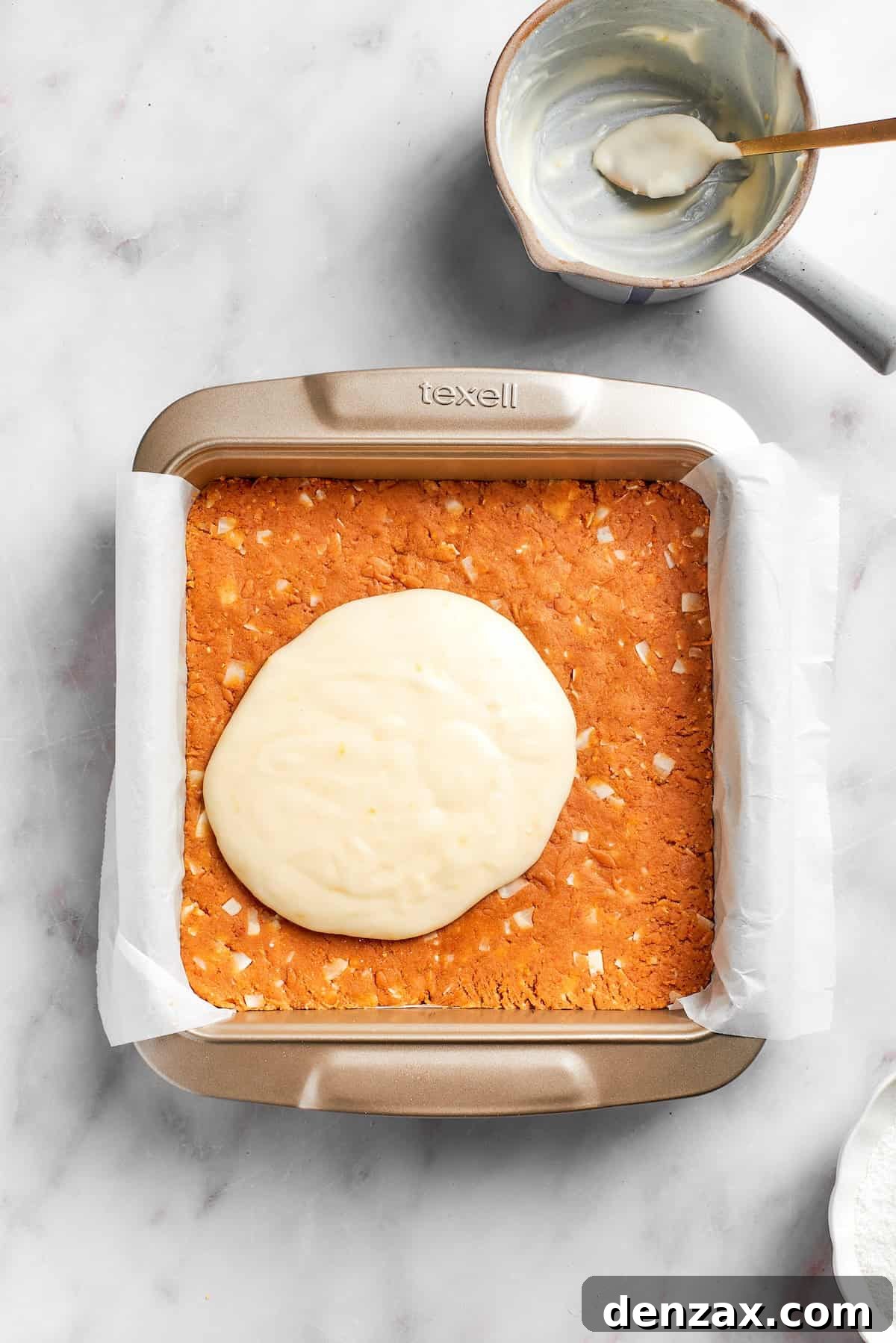 The creamy lemon frosting is generously spread over the chilled graham cracker crust in the lined baking pan, creating a smooth, even top layer.