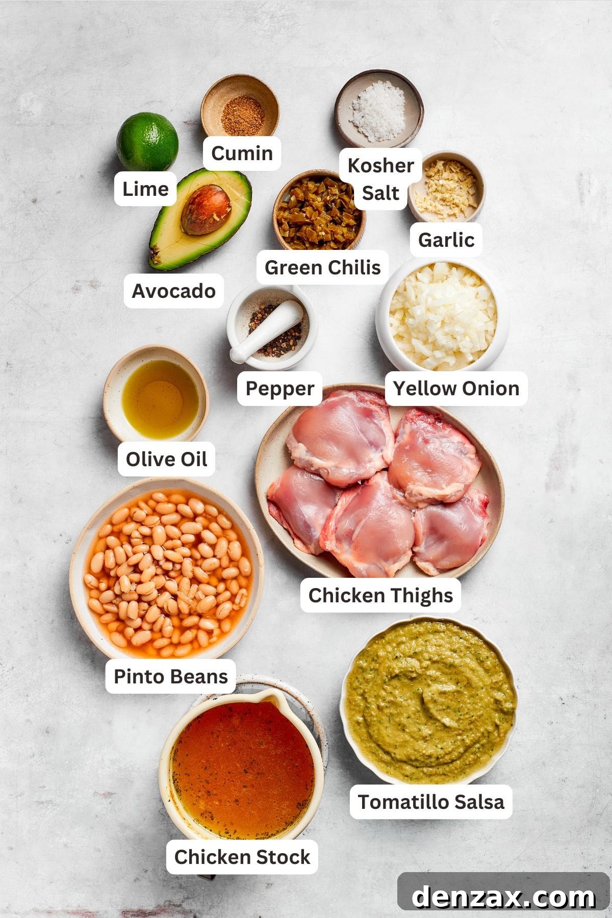 All the essential ingredients for making spicy chicken soup are laid out and clearly labeled on a rustic wooden surface, including chicken thighs, various spices, fresh vegetables, canned beans, and a jar of salsa verde, showcasing the fresh and vibrant components of the dish.