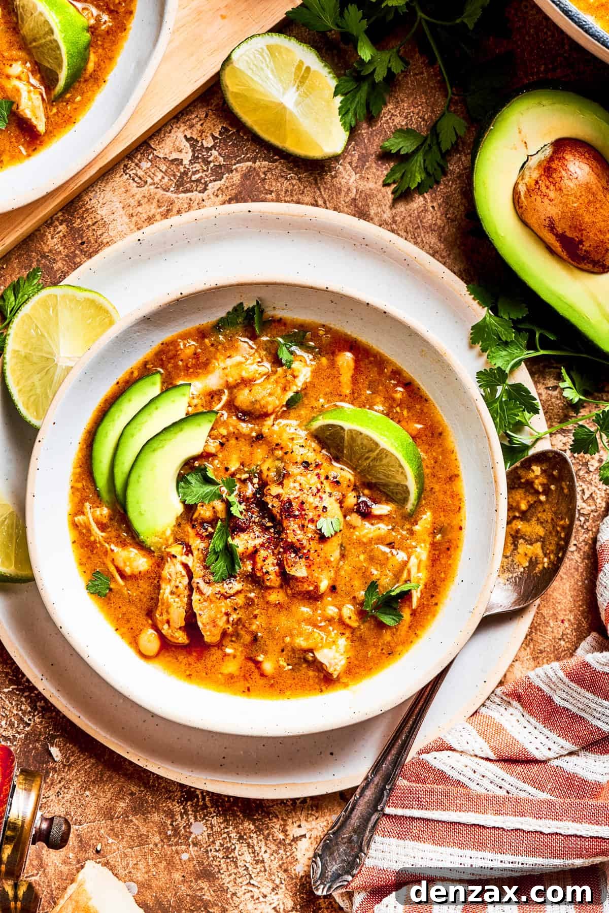A steaming bowl of spicy chicken soup, garnished with vibrant green avocado slices, fresh cilantro, and a wedge of lime, resting on a textured gray background, promising a rich and zesty flavor experience.