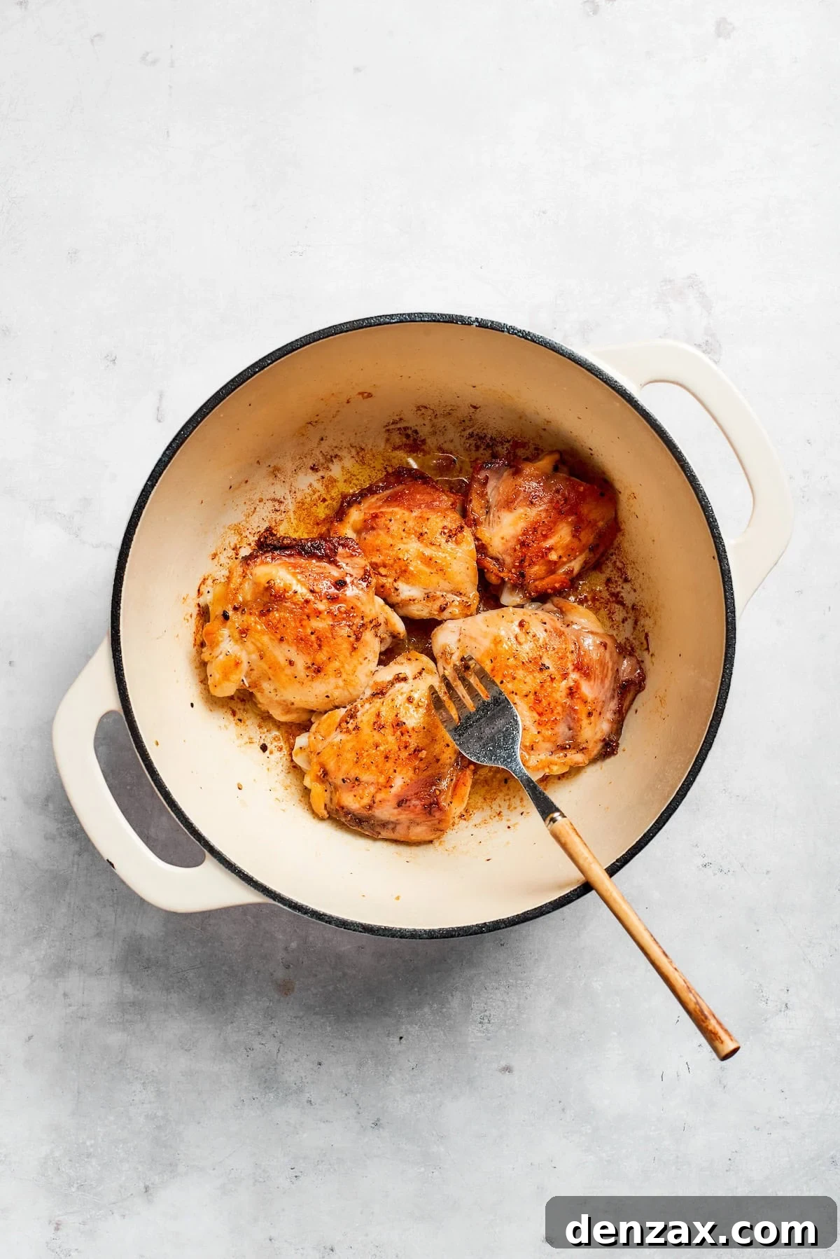 A fork lifts a piece of browned chicken from a large pot, showing the perfectly seared exterior of the chicken thigh meat. The pot contains residual olive oil and seasonings, ready for the next step.