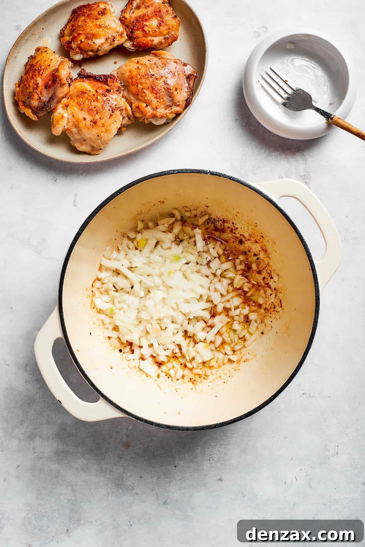 Finely diced yellow onion and minced garlic are being added to a hot pot with sizzling oil, signaling the start of building the soup's aromatic base. The pot has a slight caramelization from browning the chicken.