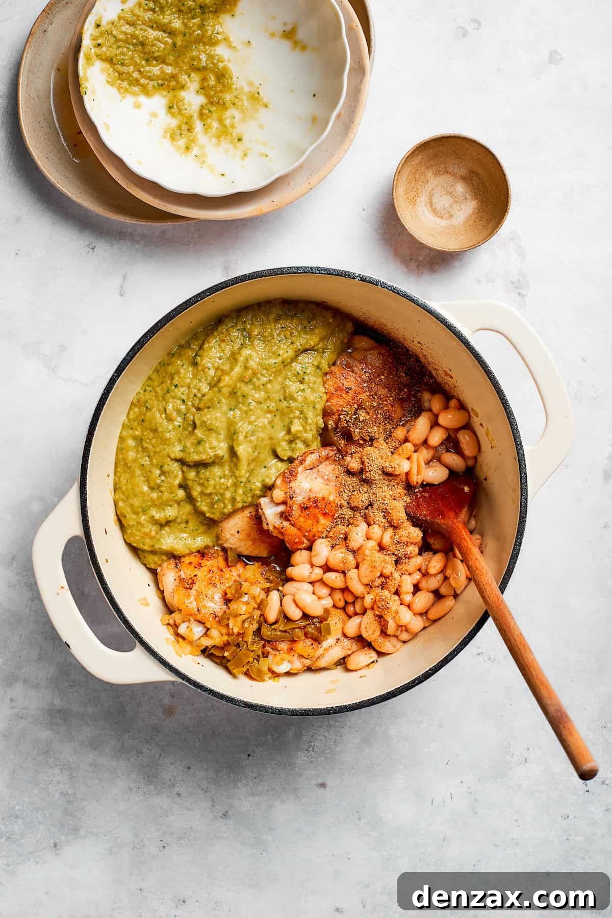 Various ingredients for the spicy chicken soup, including rinsed pinto beans, diced green chiles, and a jar of salsa verde, are being added into a large pot with sautéed onions and garlic, preparing to form the flavorful broth.
