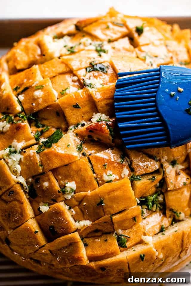 A hand brushing melted garlic butter and parsley into the scored crevices of a round loaf of bread.