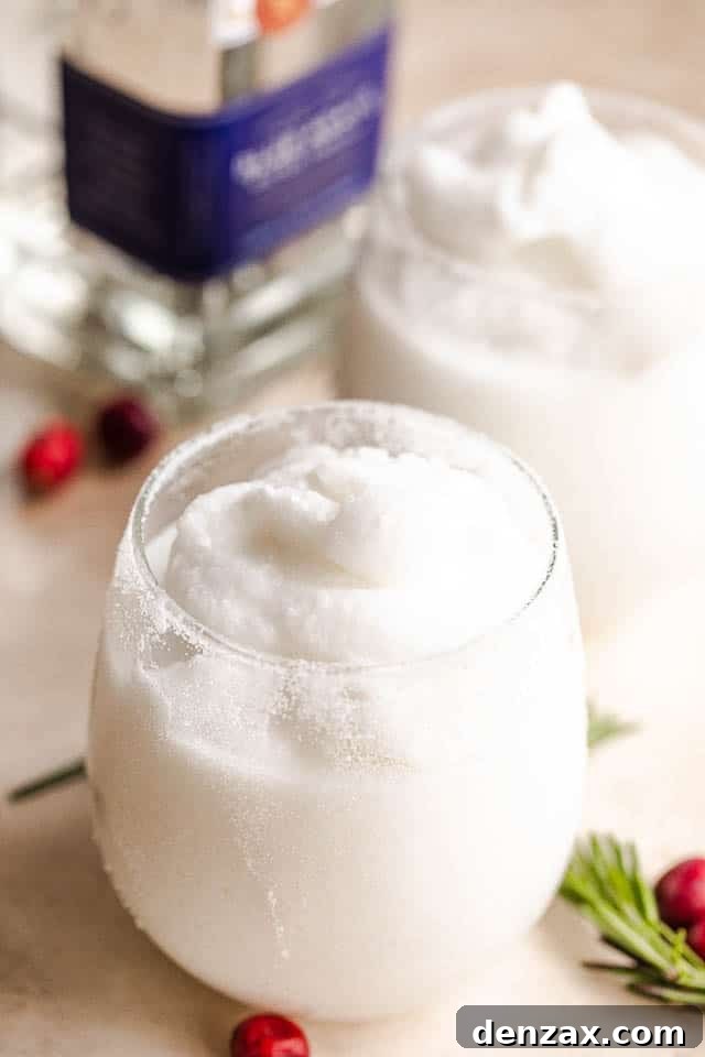 Two white frozen coconut margaritas in drinking glasses, garnished with lime and cranberries, with a bottle of silver tequila blurred in the background, on a rustic wooden surface.