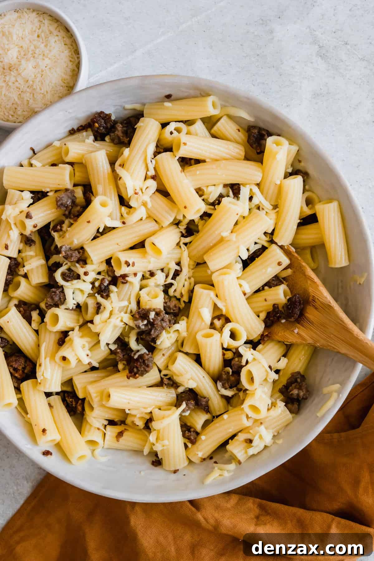 A mixing bowl containing cooked rigatoni pasta, shredded mozzarella and bits of browned ground beef, all combined and ready for the sauce.