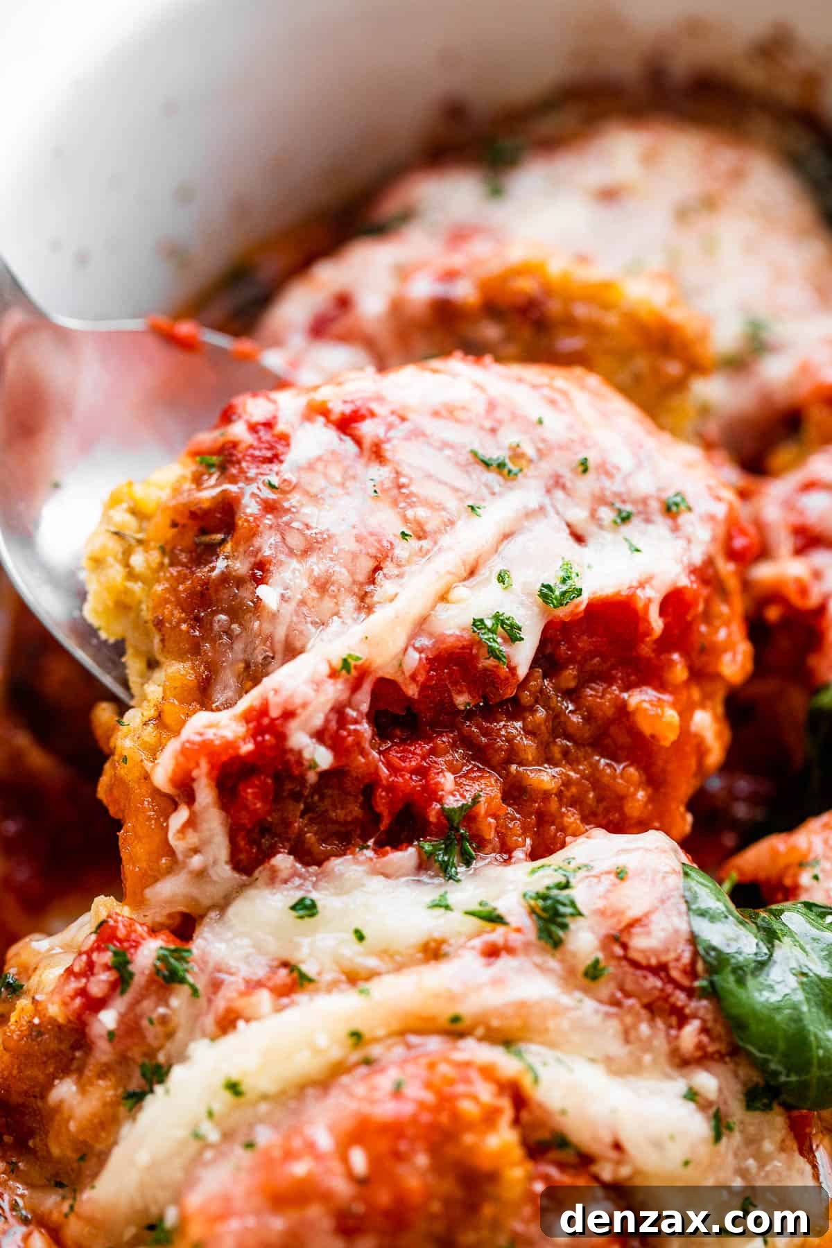 A serving spoon lifting out a baked pork parmigiana medallion from a white baking dish, showing the gooey cheese and rich sauce.