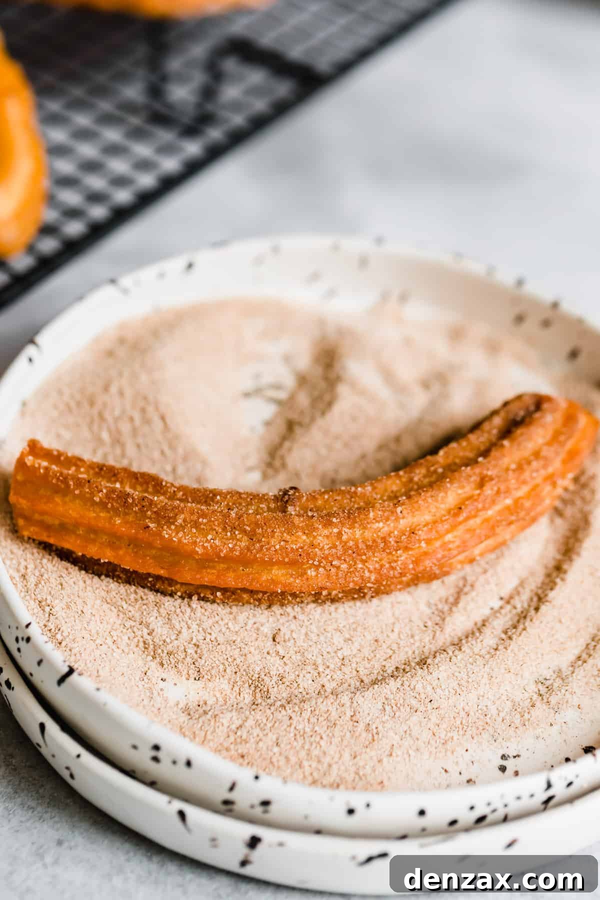 A single churro being rolled in a shallow bowl filled with aromatic cinnamon sugar, ensuring full coverage.