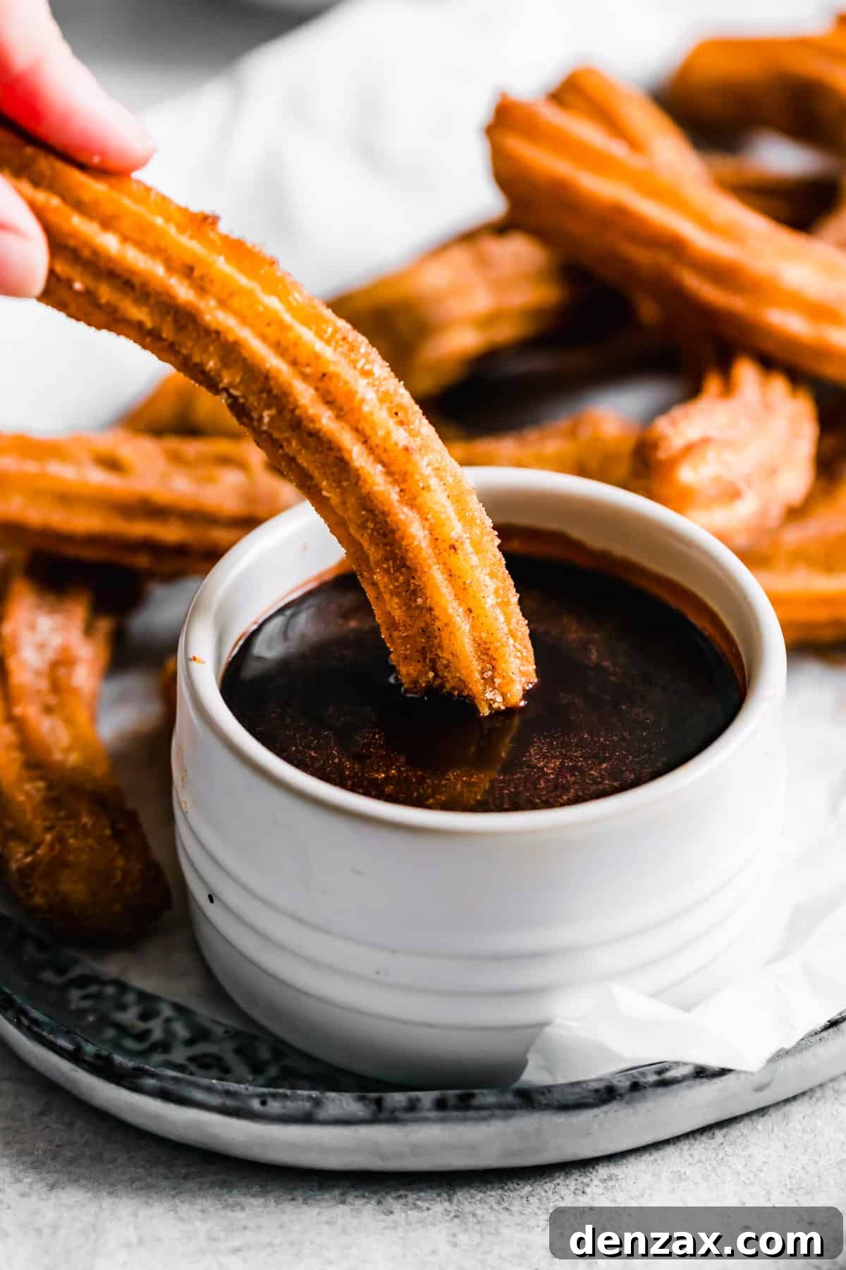 A hand dipping a fresh churro into a bowl of rich, melted chocolate sauce. Recipe by Easy Weeknight Recipes.