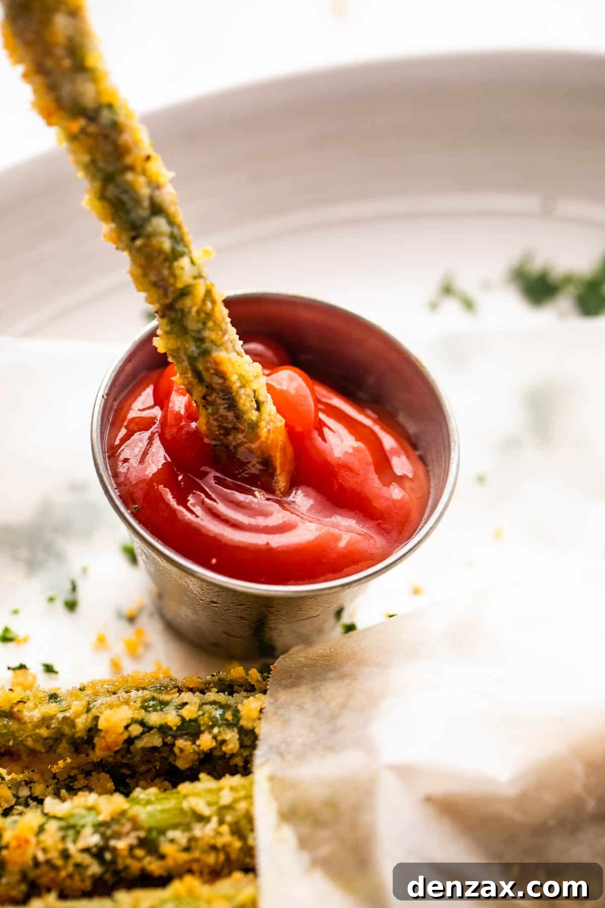 Close-up of a hand dipping a crispy baked asparagus fry into a small bowl of ketchup, highlighting the golden breading.