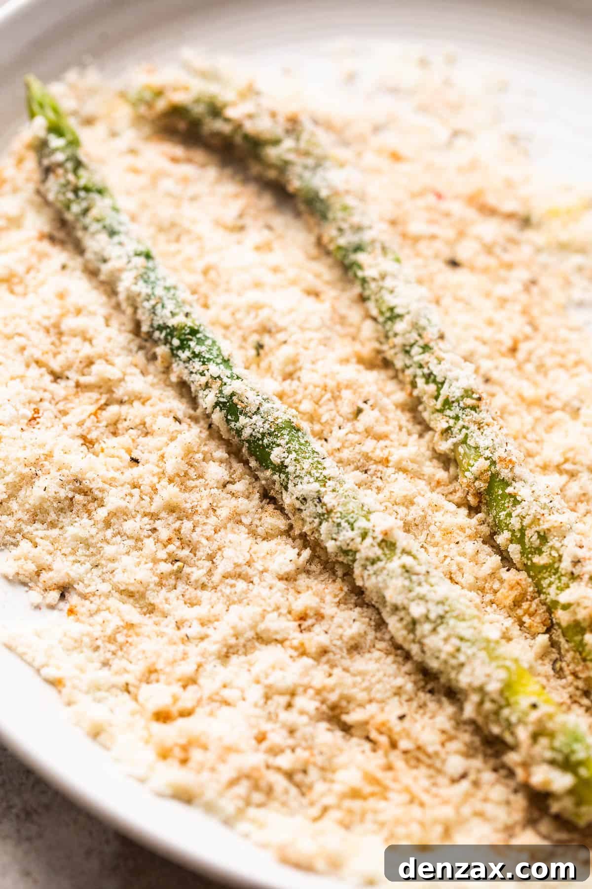 Close-up shot of fresh asparagus spears being rolled in a bowl filled with parmesan panko bread crumb mixture, ensuring a thorough coating.