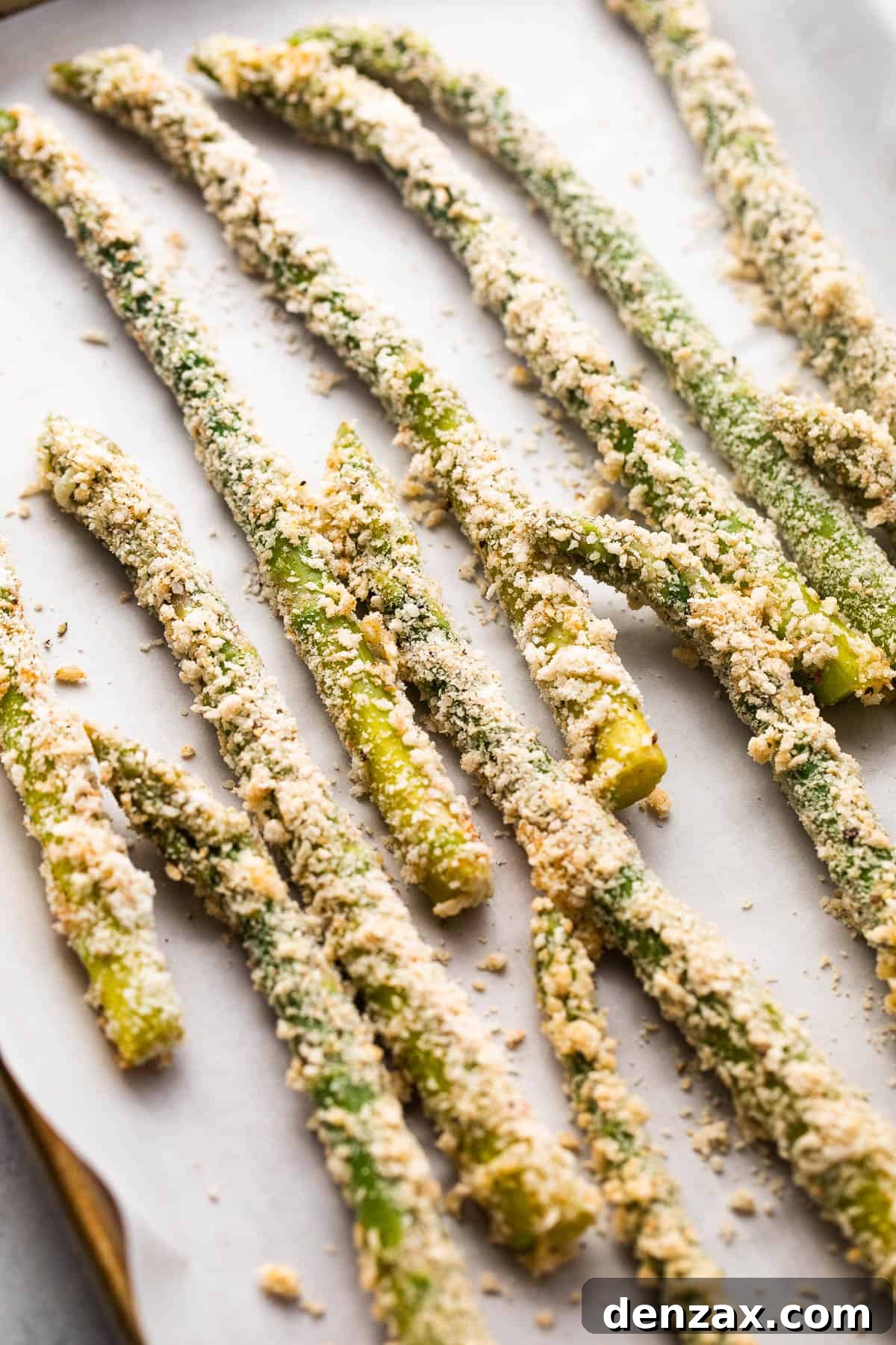 Perfectly arranged fresh asparagus spears, uniformly coated with parmesan panko crumbs, laid out on parchment paper on a baking sheet, ready for baking.