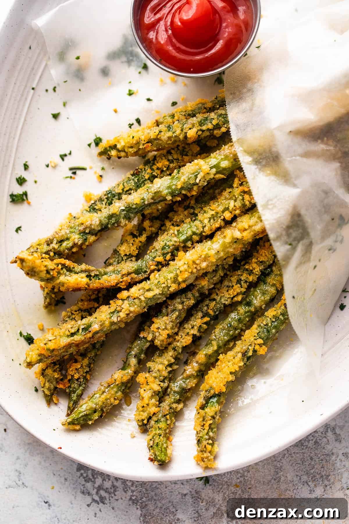 Another overhead shot of perfectly golden-brown baked parmesan panko asparagus fries served on a white plate with a side of red ketchup for dipping.