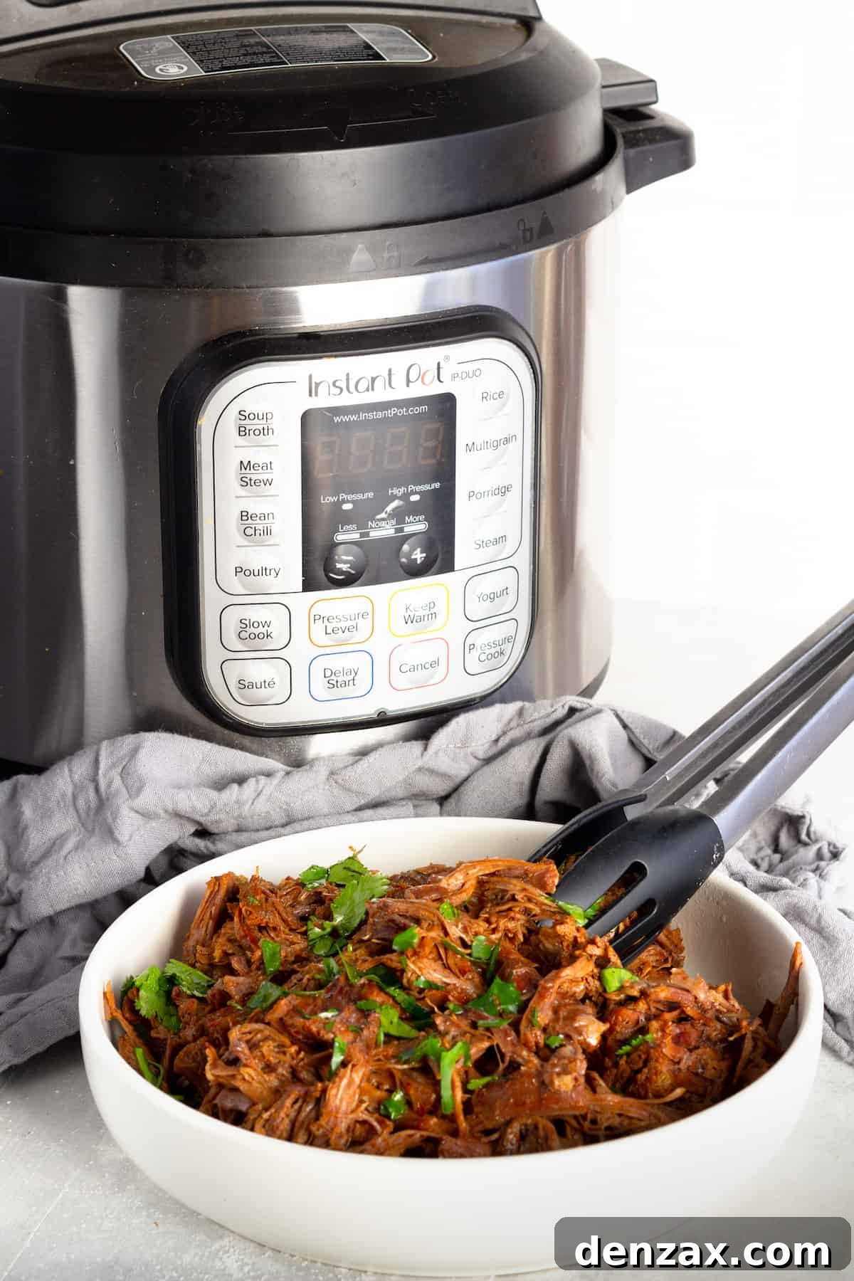 A steaming bowl of Instant Pot Beef Barbacoa sits in front of the Instant Pot itself and a cozy gray kitchen towel, highlighting the convenience of this dish.