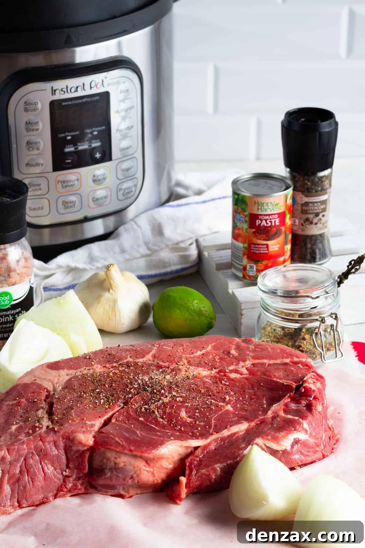 A selection of fresh ingredients for Instant Pot Beef Barbacoa, including an Instant Pot, a tube of tomato paste, a beef chuck roast, chopped onions, a fresh lime, and an array of spices, neatly arranged on a counter.