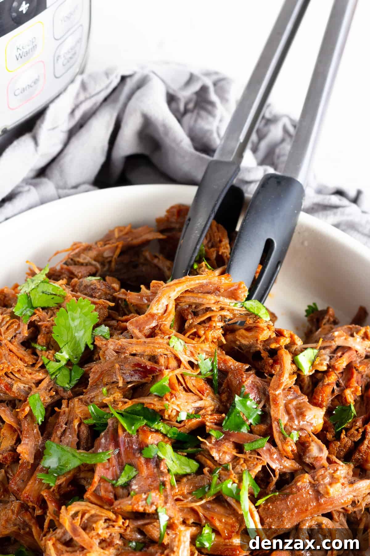 A pair of metal tongs effortlessly pulling apart tender strands of Instant Pot Beef Barbacoa in a white bowl, showcasing its perfect shreddable texture.