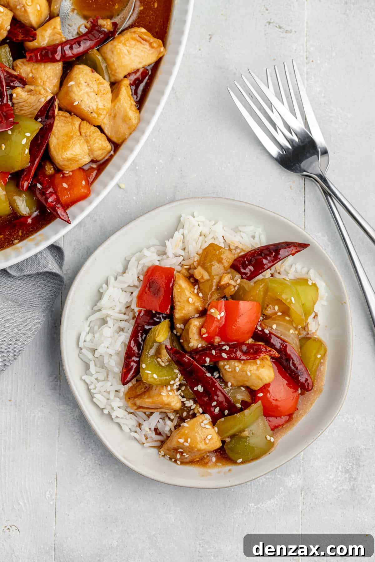 A serving of Szechuan chicken stir-fry over rice with a serving platter and two forks nearby on the table, invitingly presented.