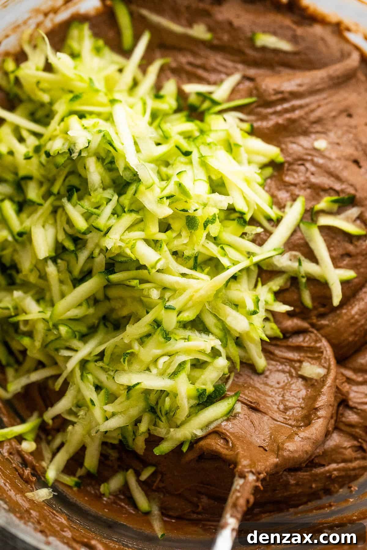 Shredded zucchini being mixed into rich chocolate batter with a spatula