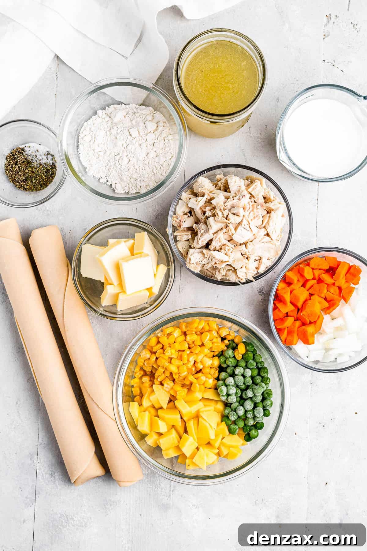 Clockwise from top: chicken broth, milk, chopped carrots and onions, a bowl of peas, corn, and diced potatoes, butter, seasonings, and flour, laid out as ingredients for a chicken pot pie.