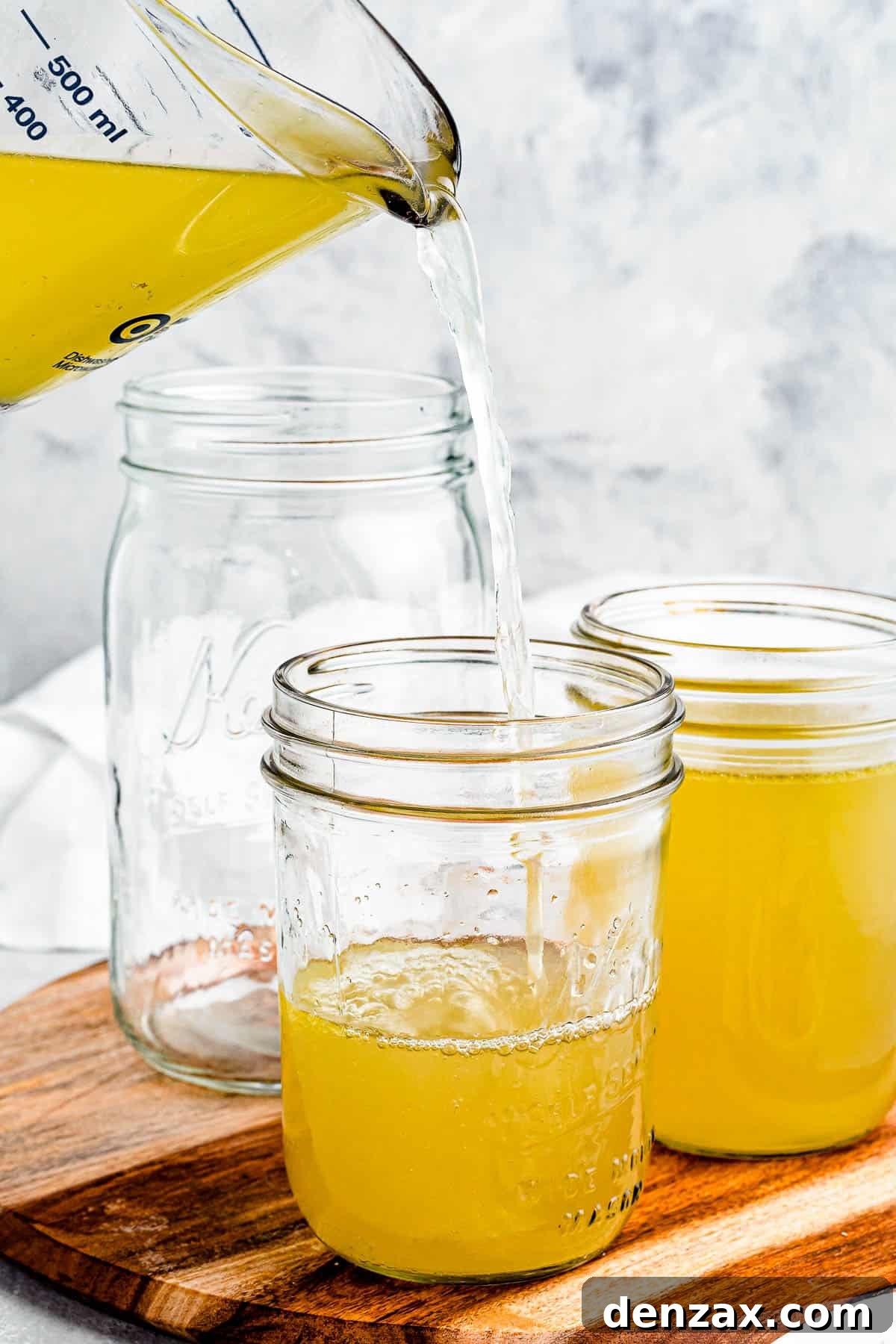 Chicken broth being poured from a large measuring cup into several jars.