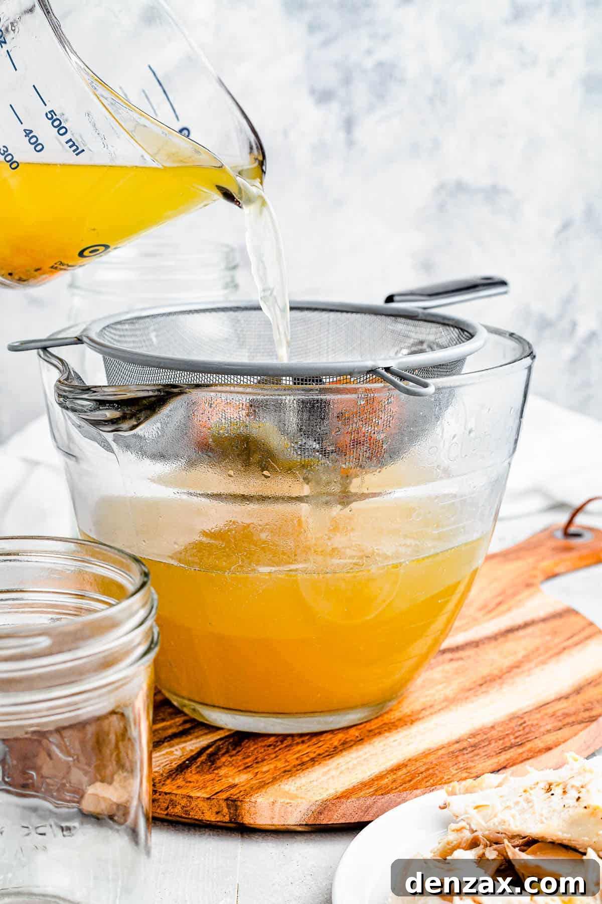 Chicken broth being poured from a large measuring cup into a bowl through a large sieve.