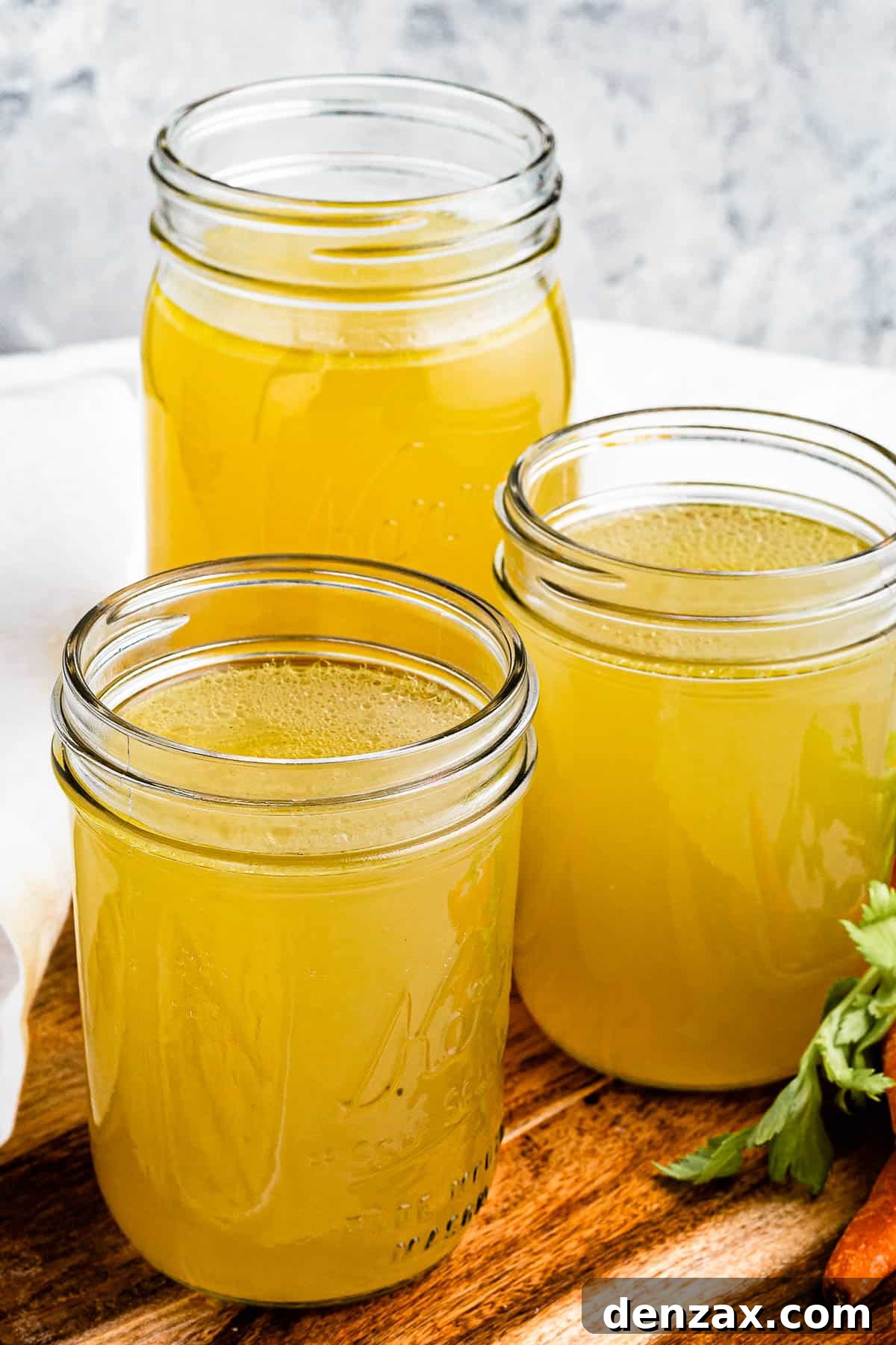 Three jars of chicken broth on a cutting board with raw carrots positioned next to them.