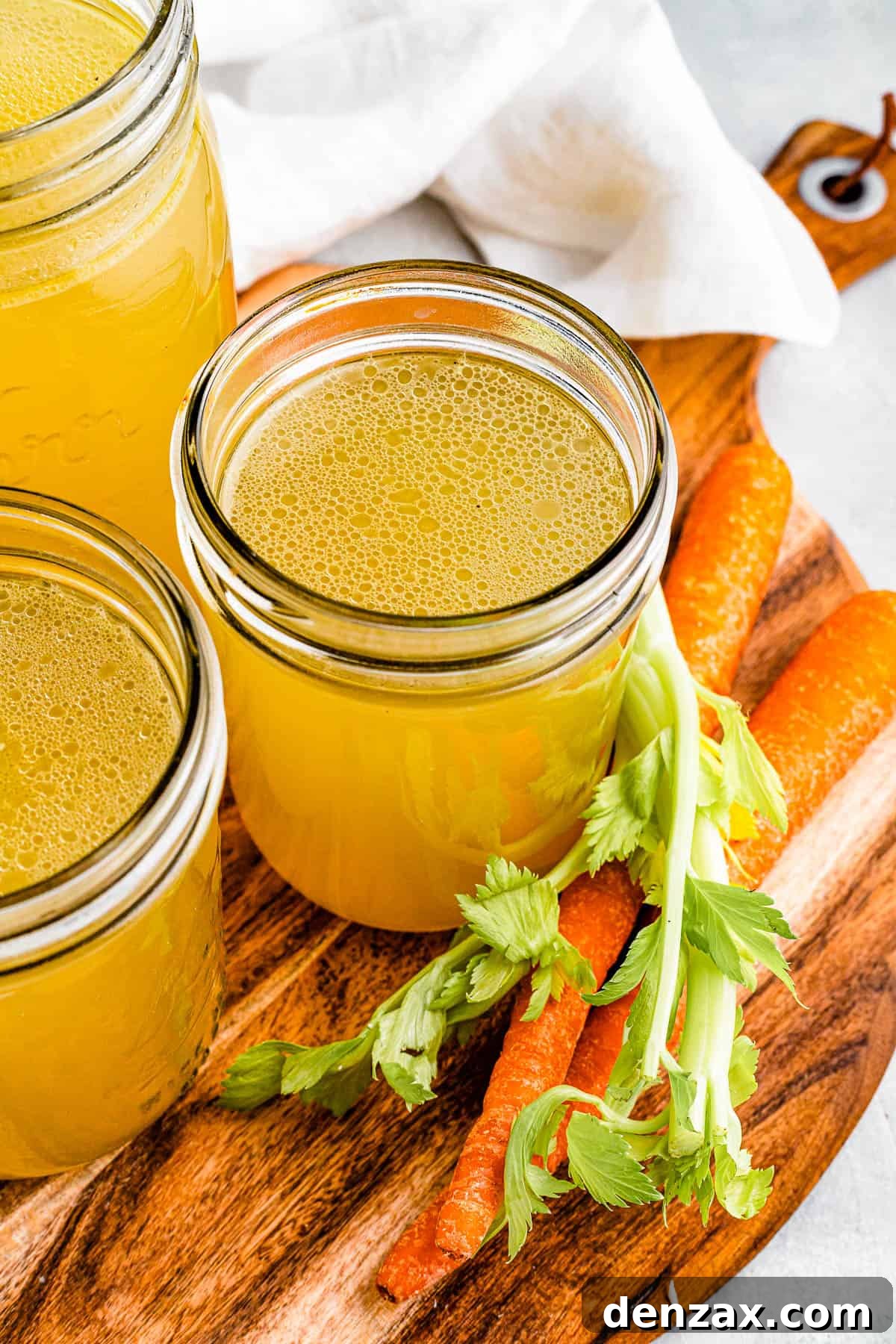 Three jars of chicken broth on a cutting board with raw carrots nearby.