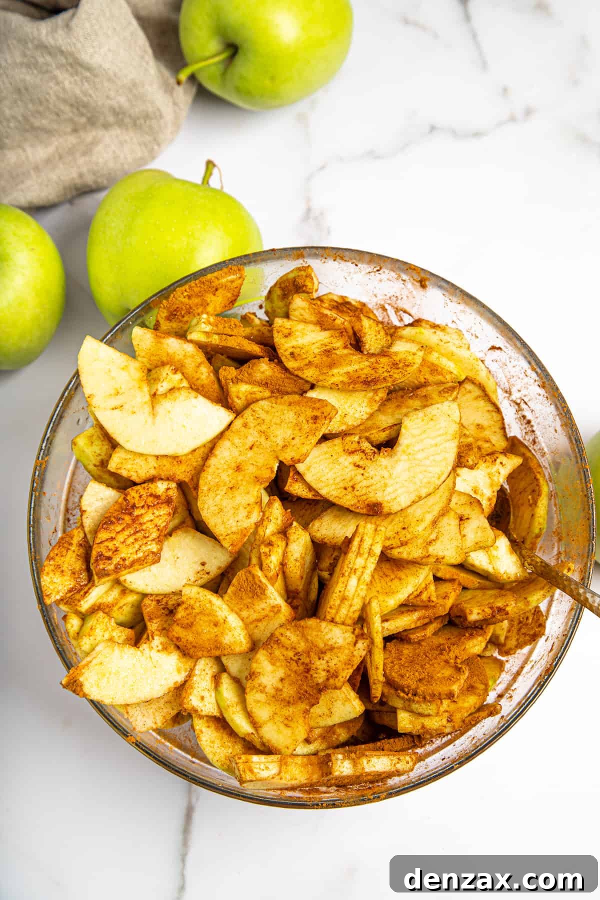 Thinly sliced apples coated thoroughly with cornstarch, sugar, and warming spices in a mixing bowl.