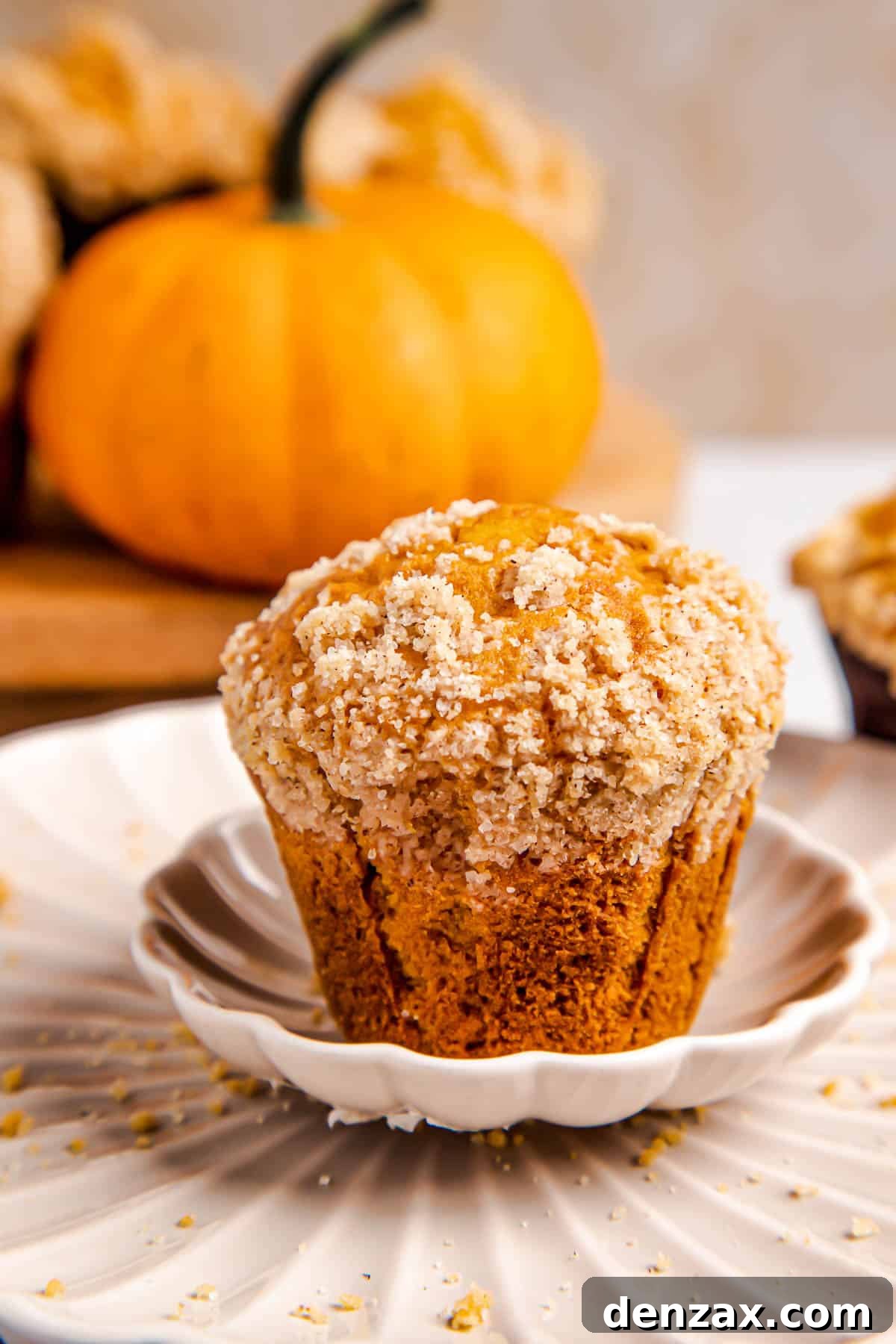 A single, perfectly baked pumpkin muffin, rich with cinnamon crumble, resting on a white plate with more muffins softly blurred in the background, hinting at the warm, inviting experience of homemade fall treats.