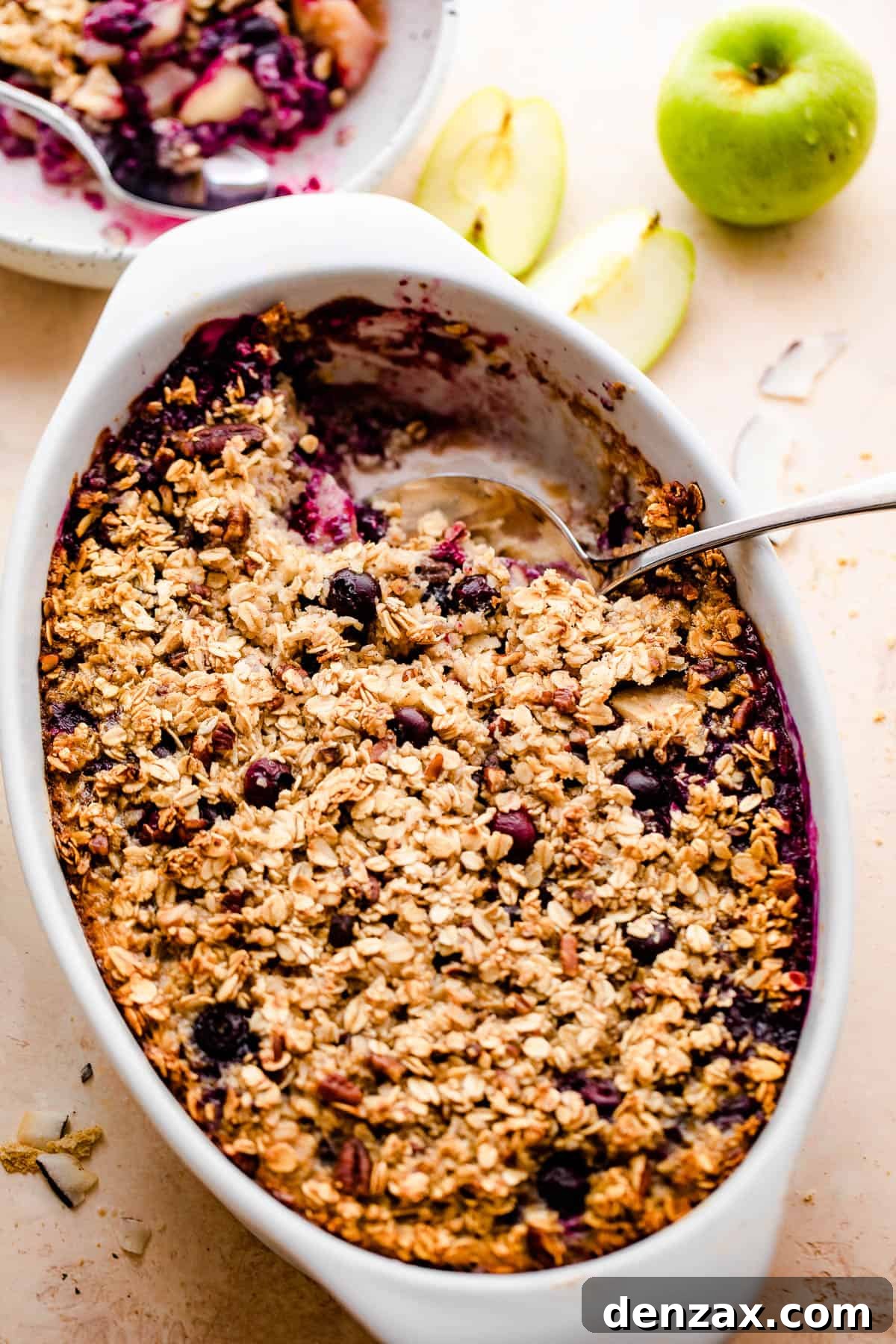 overhead shot of baked oatmeal in a white oval baking dish with a green apple set on the side, and a plate with oatmeal right above it. 