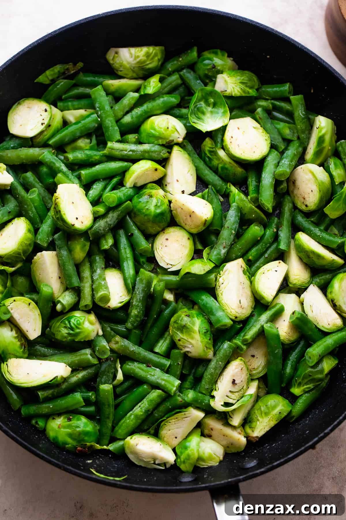 Close-up of Brussels sprouts and green beans gently sautéing in a black skillet, glistening with oil.