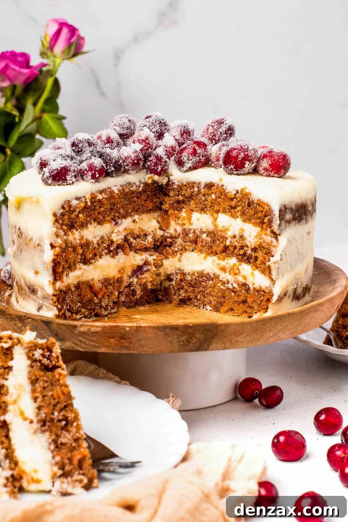 A three-layer carrot cake on a cake stand, with two slices cut out and set on dessert plates nearby. A vase of roses adds to the festive background.