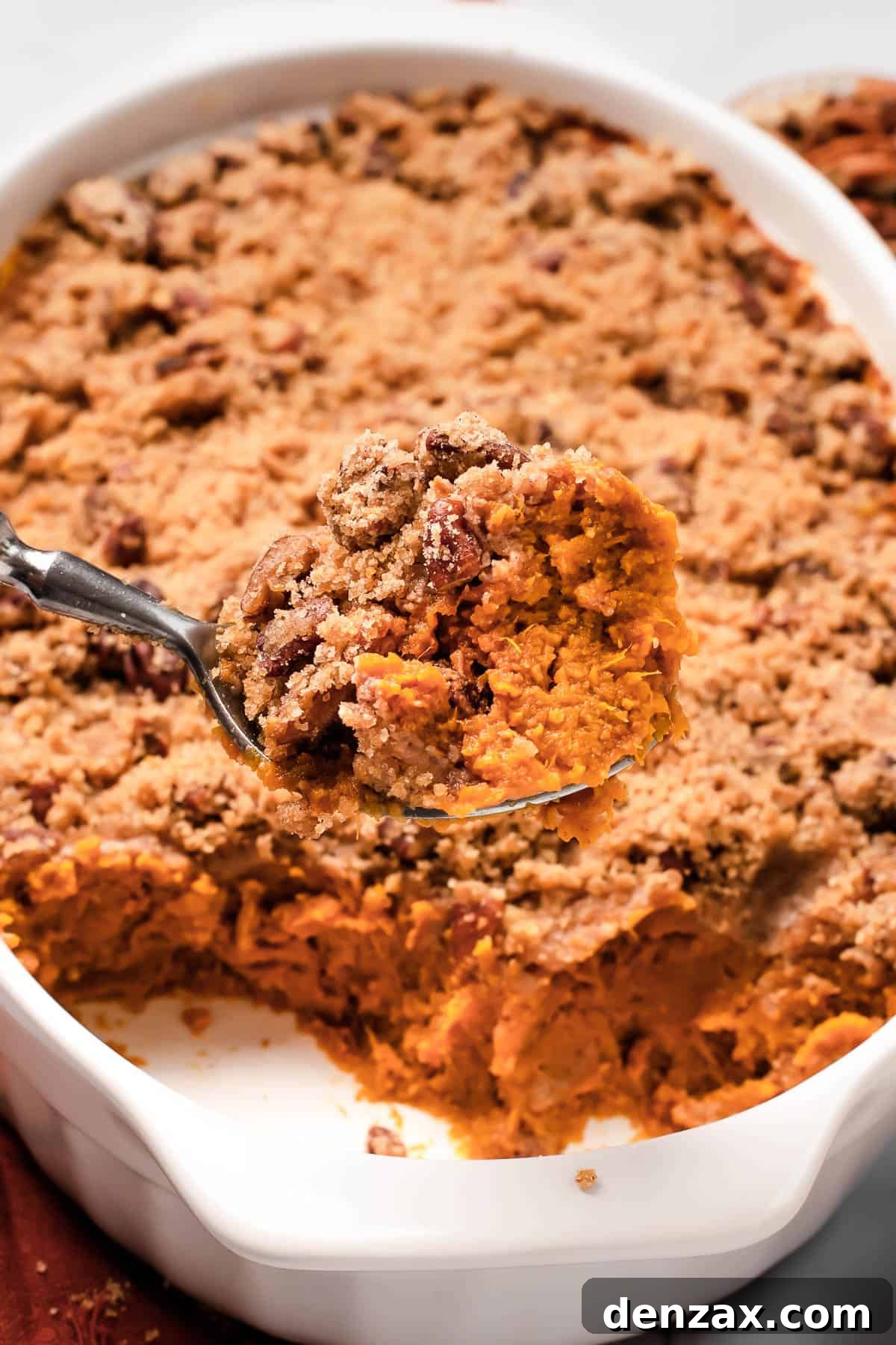 A generous serving of Sweet Potato Casserole being lifted from a baking dish with a spoon, showcasing its creamy interior and golden-brown, crunchy pecan streusel topping.