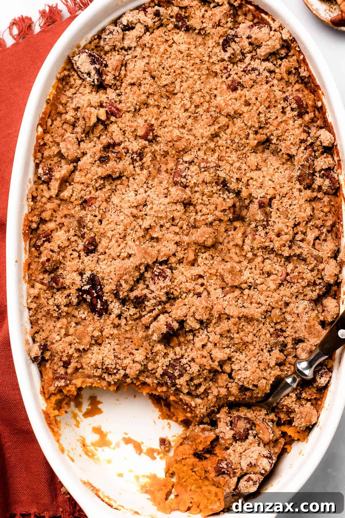 A beautifully baked dish of Sweet Potato Casserole with a golden pecan streusel topping, artfully arranged on a marble countertop with a serving spoon resting beside it.