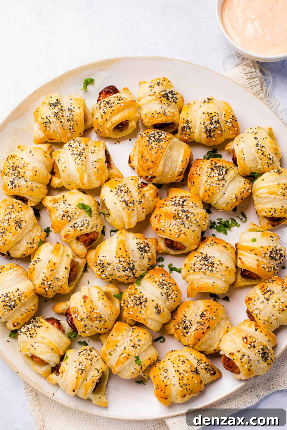 A serving platter holding about two dozen pigs in a blanket. A bowl of dipping sauce sits nearby.