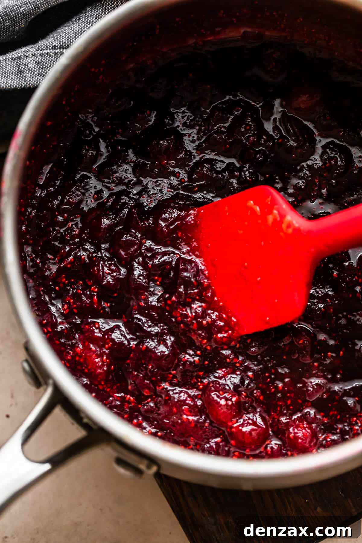 Homemade cranberry sauce simmering gently in a saucepan, being stirred with a red rubber spatula, showcasing its rich texture.