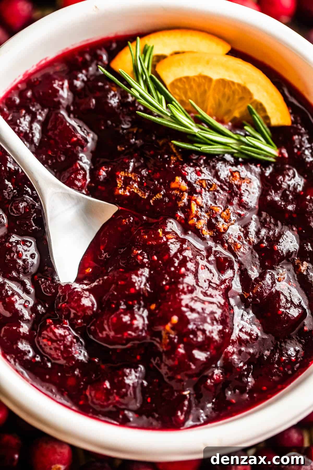 Overhead close-up shot of a spoon resting inside a bowl of homemade cranberry sauce, garnished with a fresh rosemary sprig and bright orange slices.