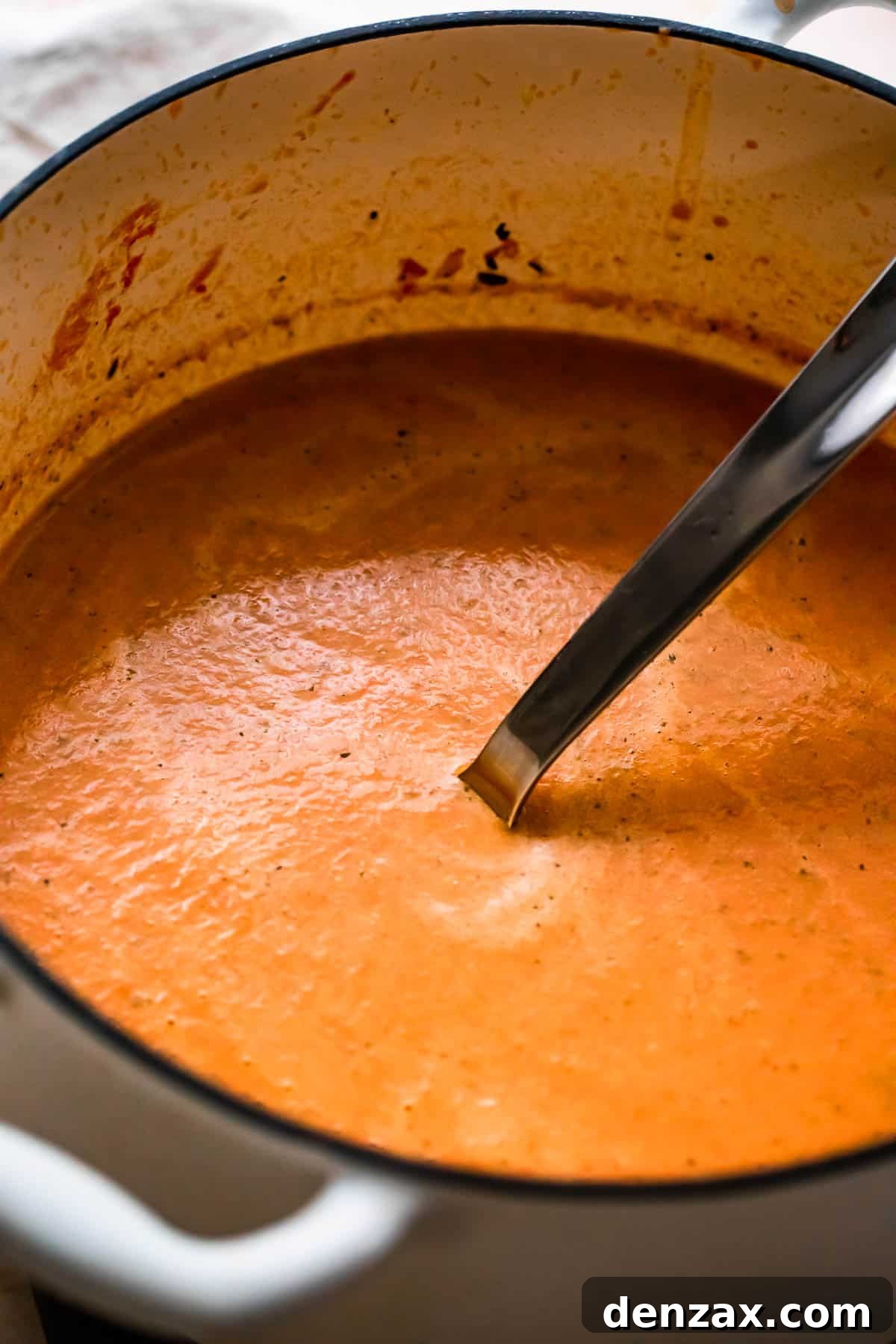 Side view of a white Dutch oven brimming with creamy roasted tomato basil soup, with a ladle resting inside.