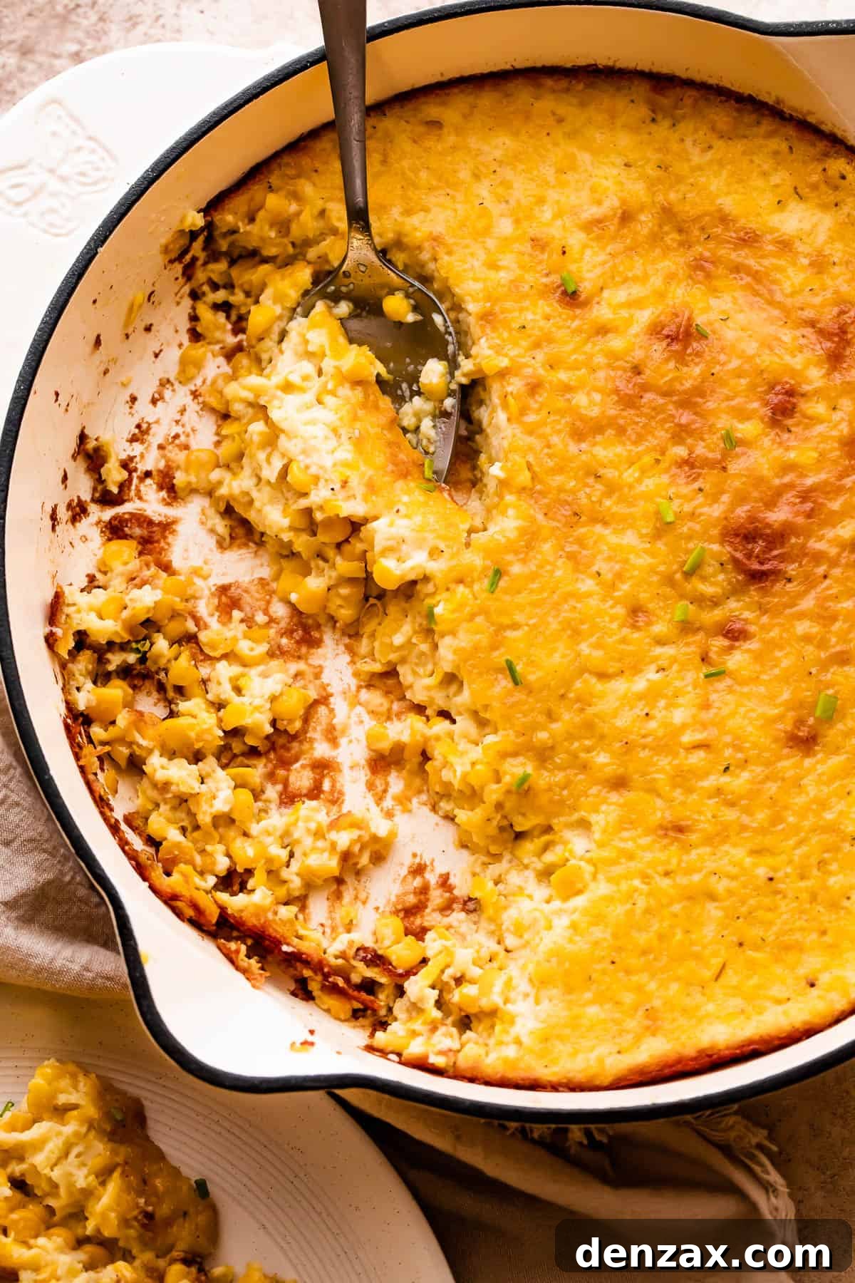 A spoon lifting a serving of creamed corn pudding from a white cast iron skillet.