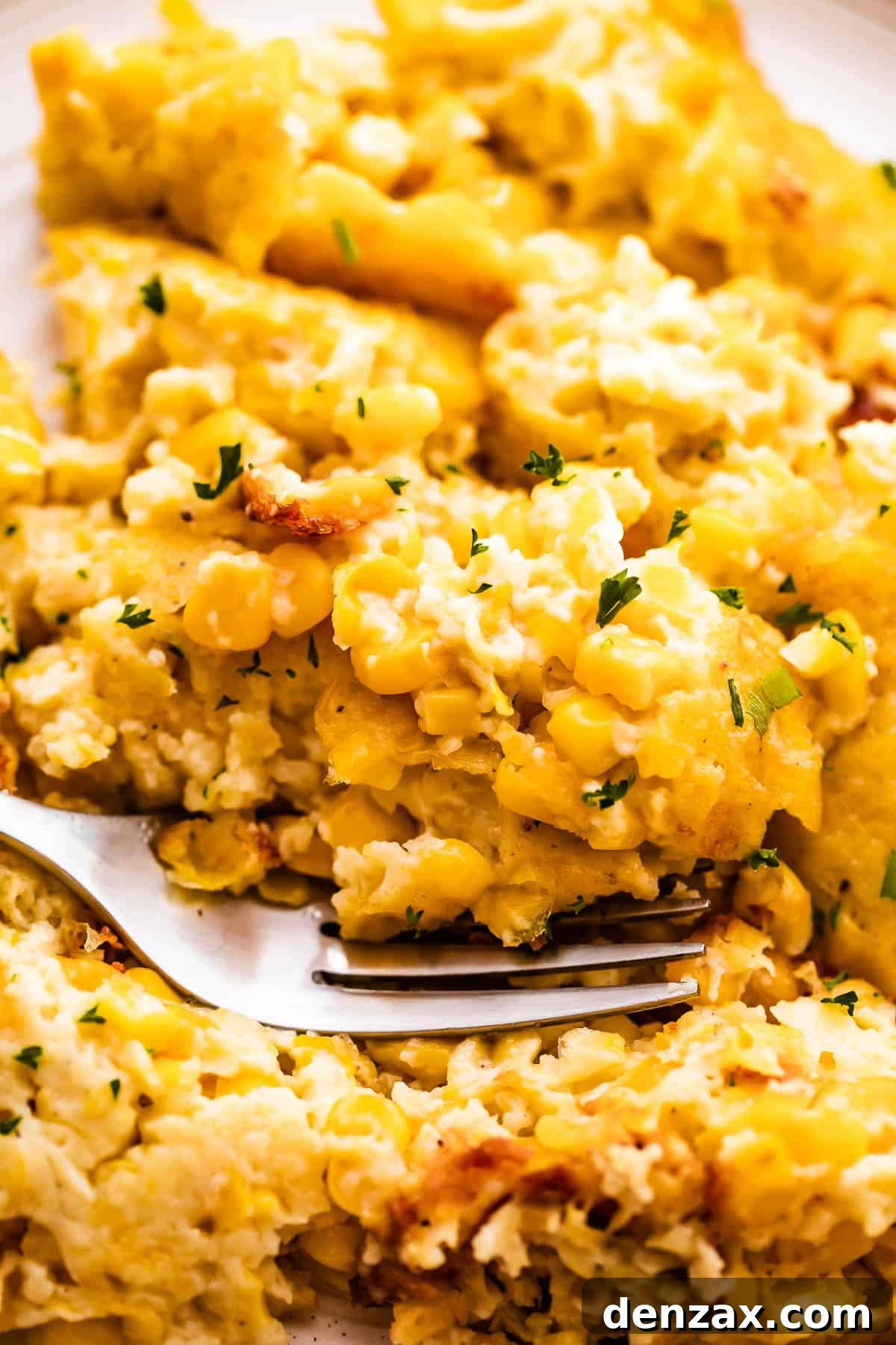 A shallow dish holding a serving of creamed corn casserole, near a baking dish of the same casserole. A spoon is in the bowl as well.