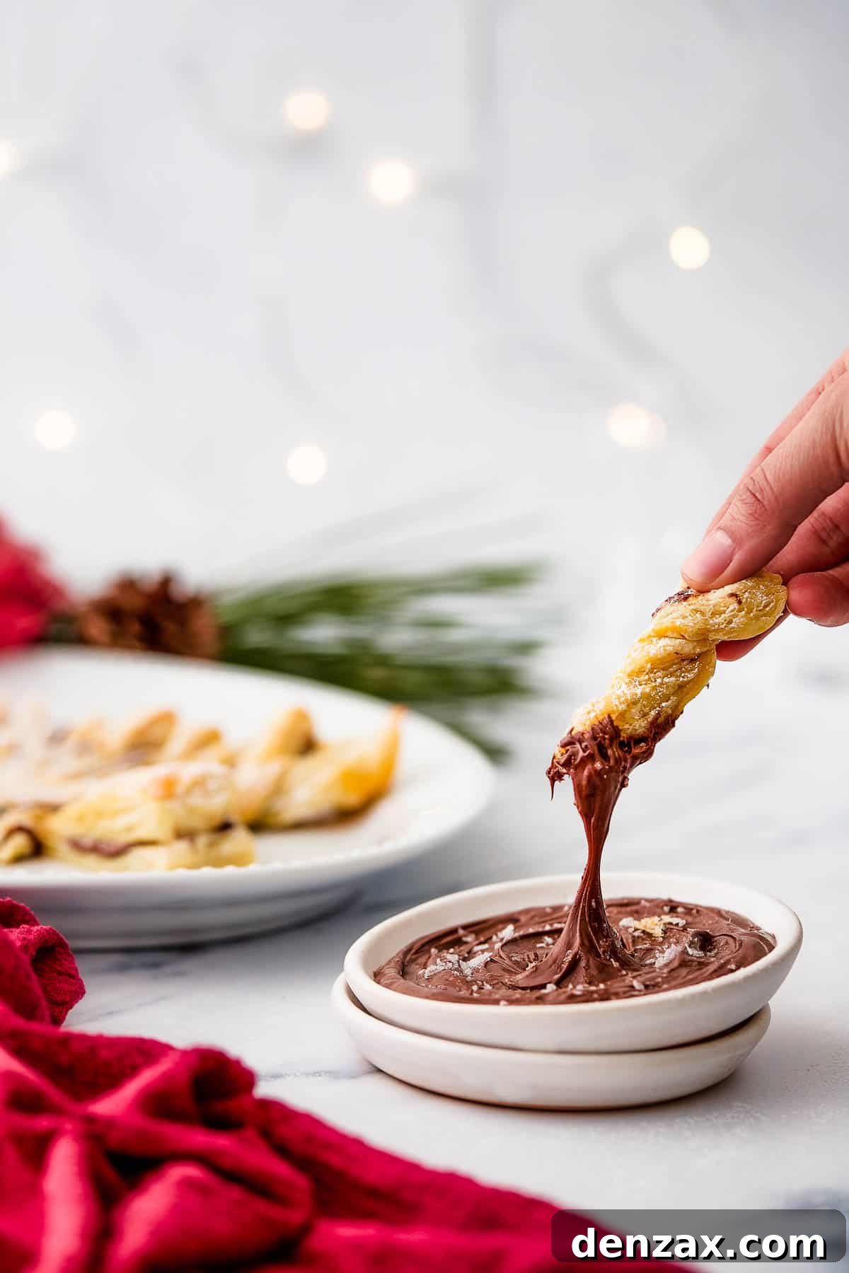A woman's hand dips a puff pastry twist into chocolate dipping sauce.Christmas decorations and a platter are in the background of this shot.